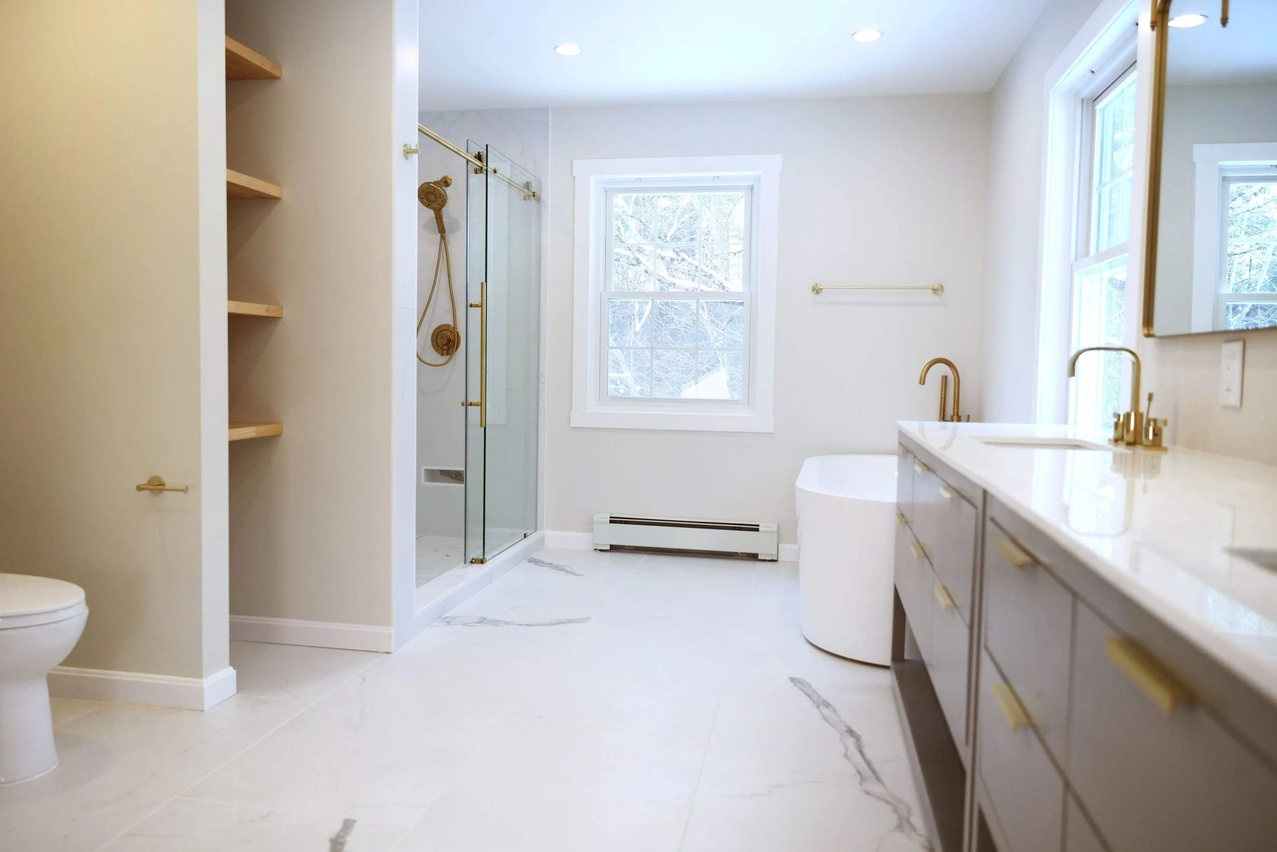 Modern bathroom with white walls, marble floor, gold fixtures, glass shower, window with snowy view, and a vanity with gold handles and a sink.