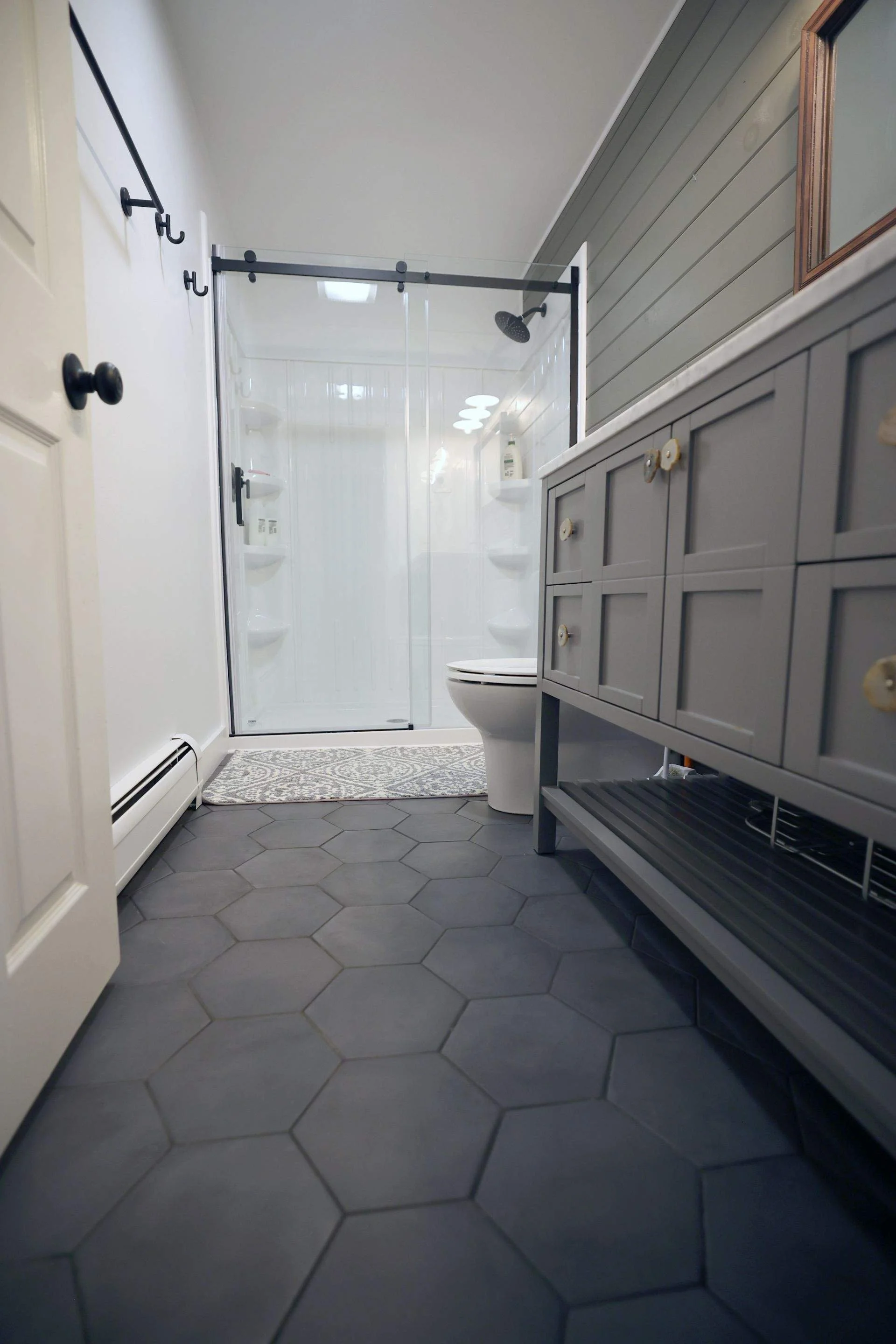 A bathroom featuring hexagonal gray floor tiles, a white toilet, a gray wooden vanity with a slatted shelf, and a glass-enclosed shower with built-in shelving and a rain showerhead.