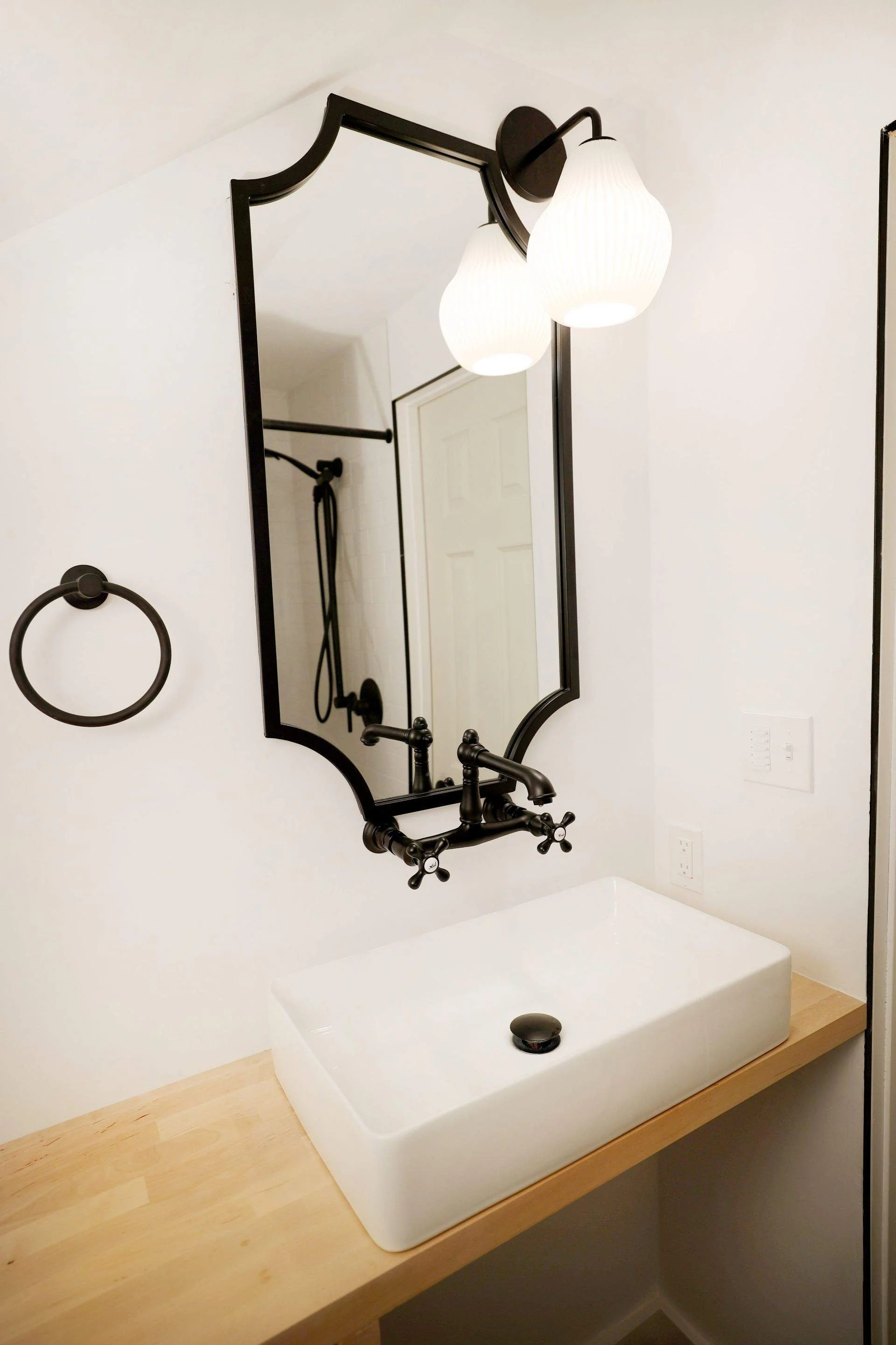 Bathroom with a rectangular white ceramic sink on a wooden countertop, a black-framed mirror with decorative edges, a black wall-mounted light with two white glass shades, a black towel ring on the wall, and a shower area with a black shower fixture and a handheld showerhead.