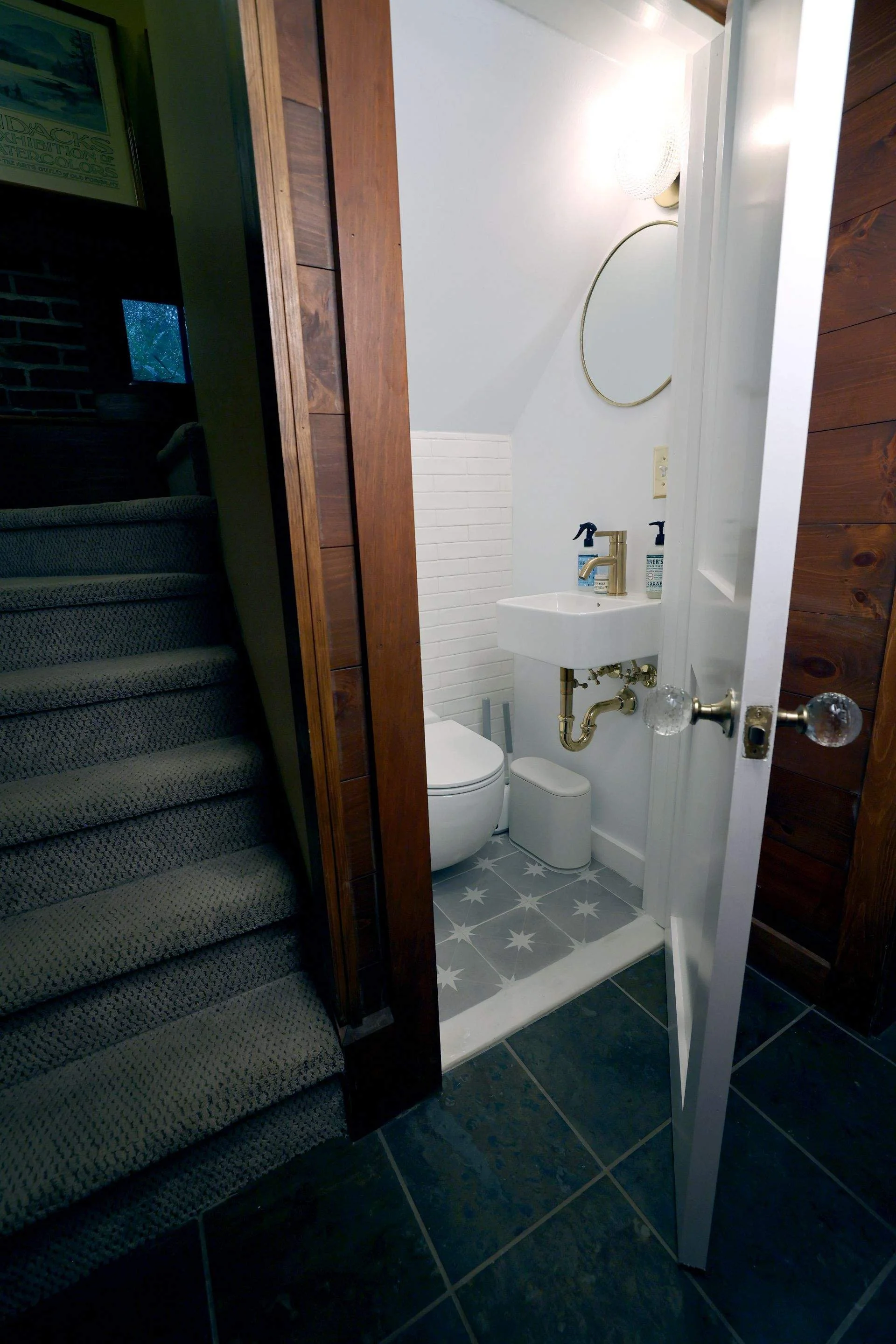 A small bathroom near a staircase with a white toilet, small sink with gold fixtures, a mirror, and patterned gray and white star tiles on the floor.