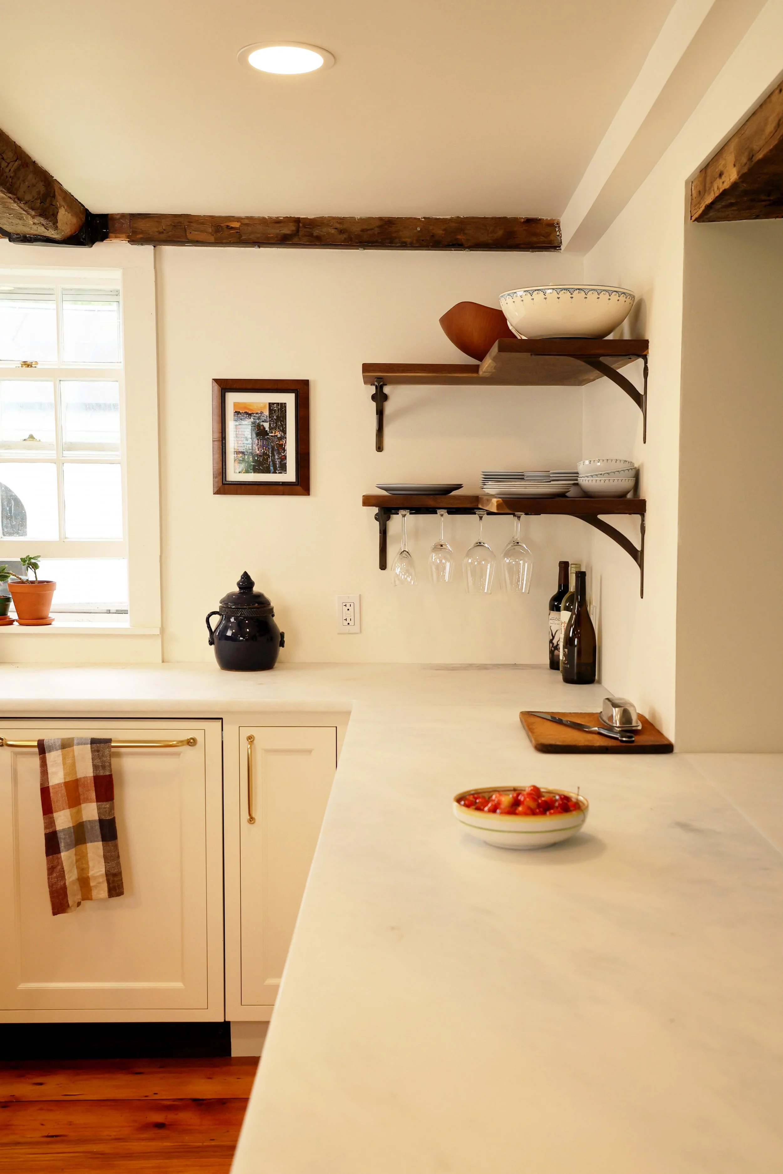 A kitchen with white cabinets, a white marble countertop, open wooden shelves holding bowls and plates, hanging wine glasses, and bottles of wine on the counter. There is a window with potted plants, a framed picture on the wall, and a bowl of cherry tomatoes on the counter.