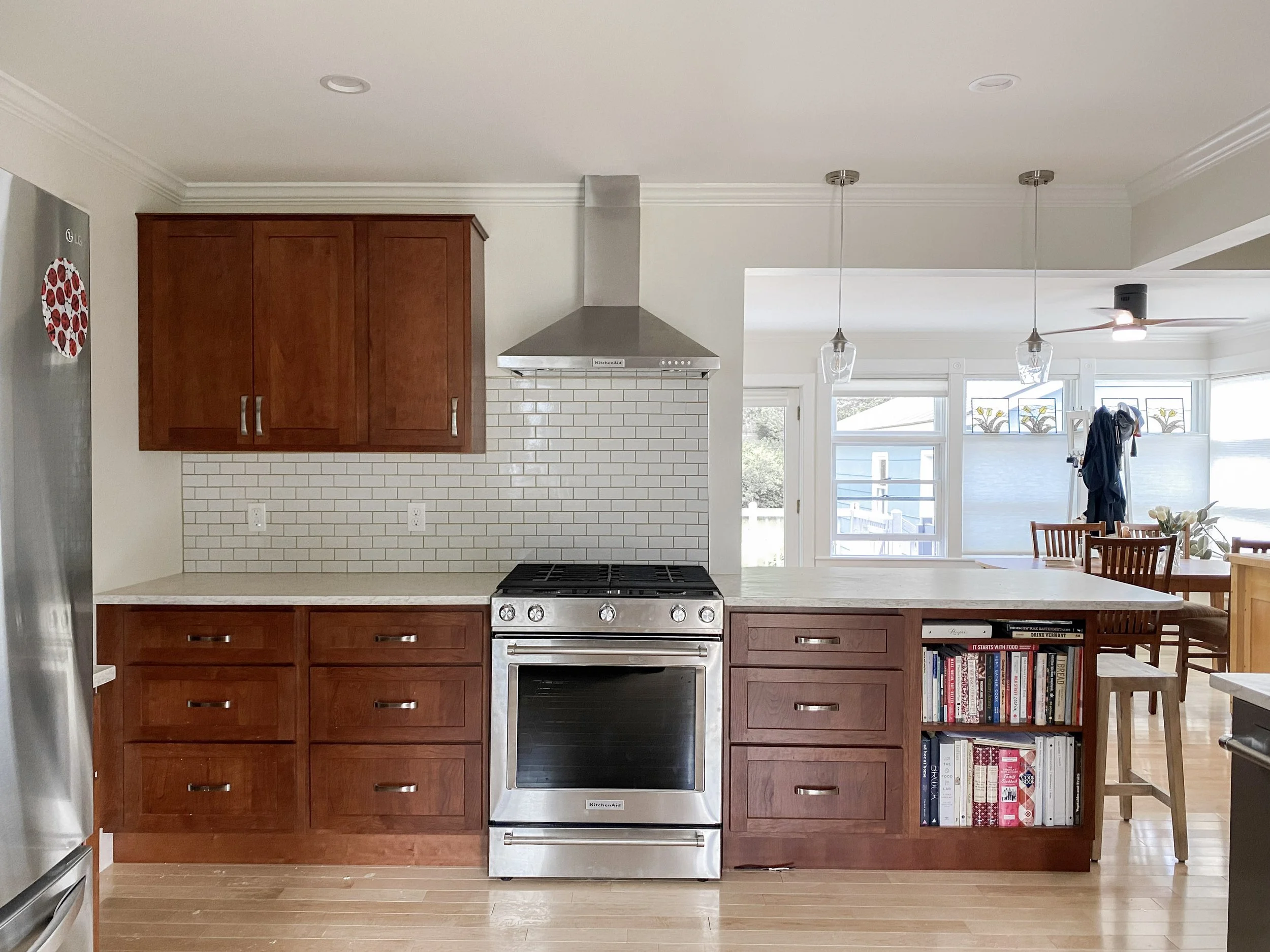 Kitchen with wooden cabinets, stainless steel stove, white subway tile backsplash, and an open bookshelf in the counter. There is also a dining area with a table and chairs and windows with blinds in the background.