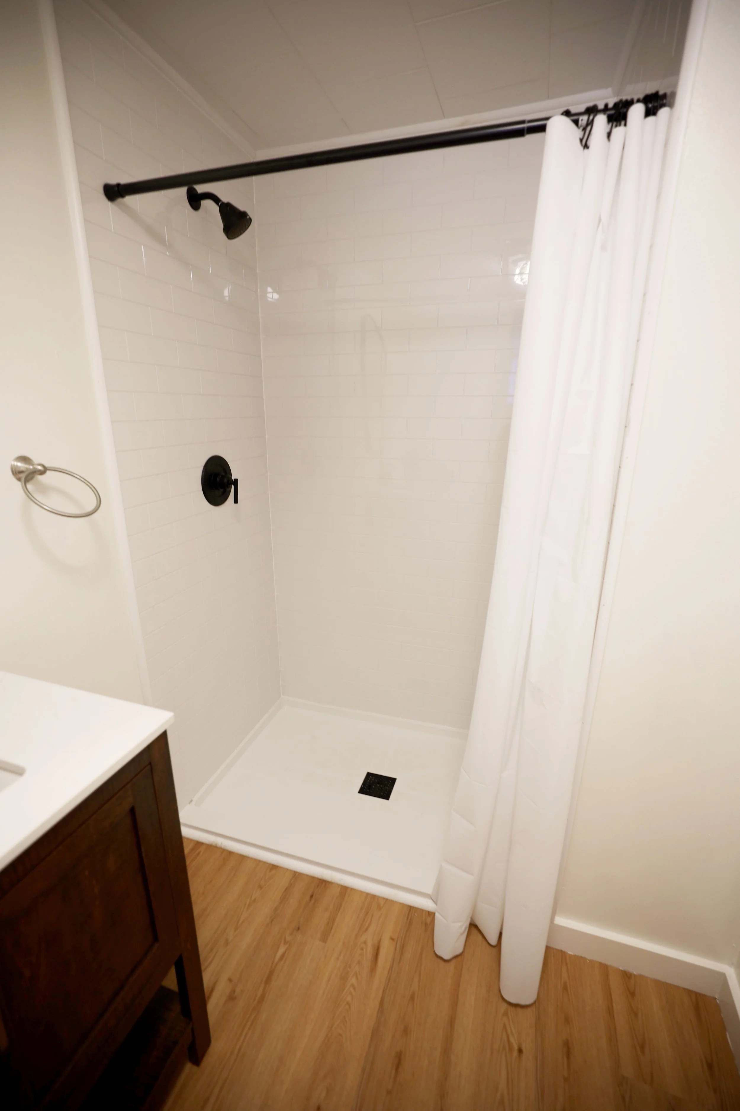 Empty shower with white tile walls, a black showerhead and control, a white curtain, and a wooden floor outside the shower.