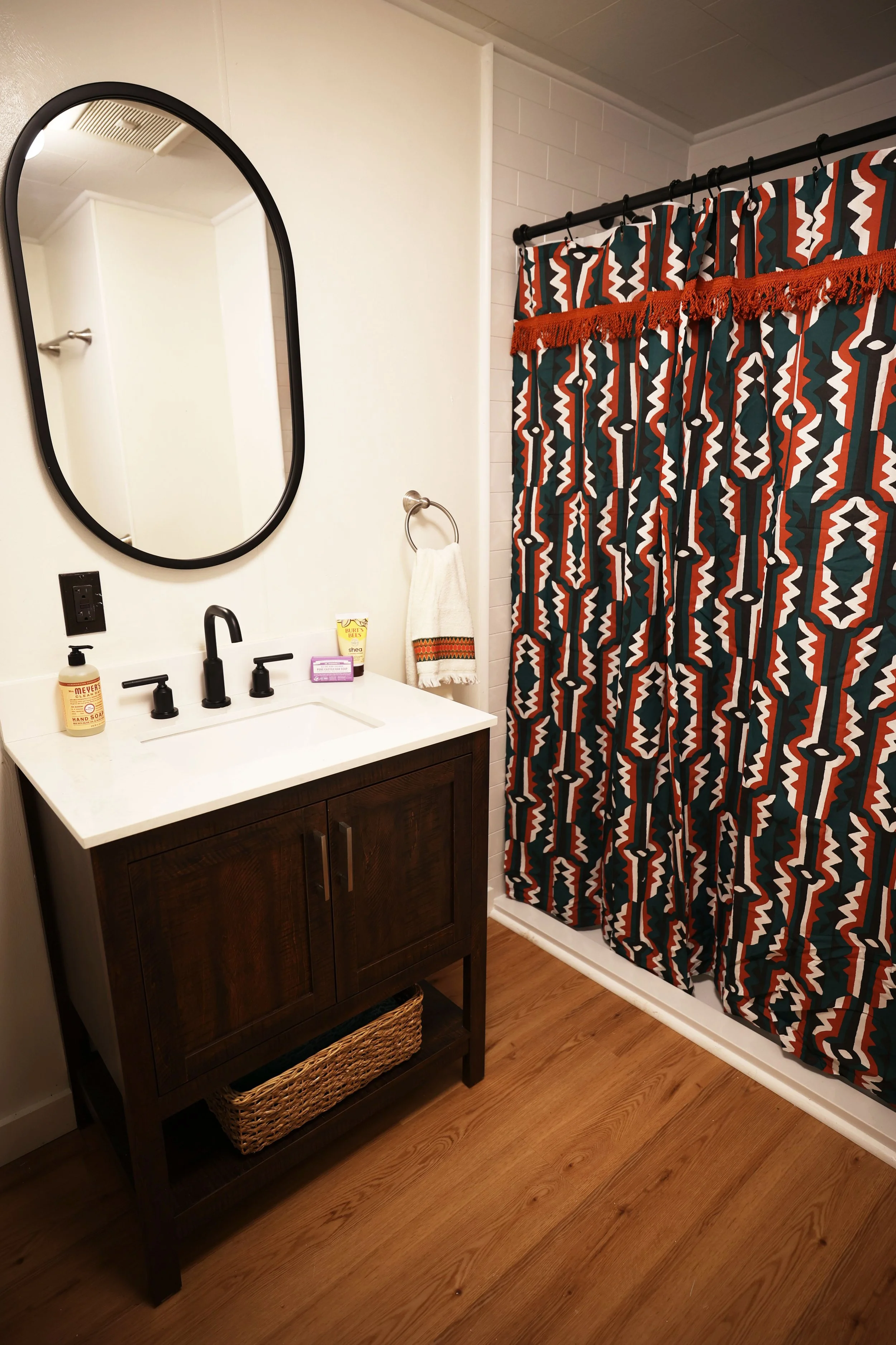 A bathroom with a dark wood vanity, a white countertop, and a black faucet. There is a mirror above the sink, a hand towel hanging on a ring, a soap dispenser, and a small container on the counter. A colorful patterned shower curtain with red, black, white, and green stands next to the vanity. The floor is wooden, and the wall behind the vanity is white.