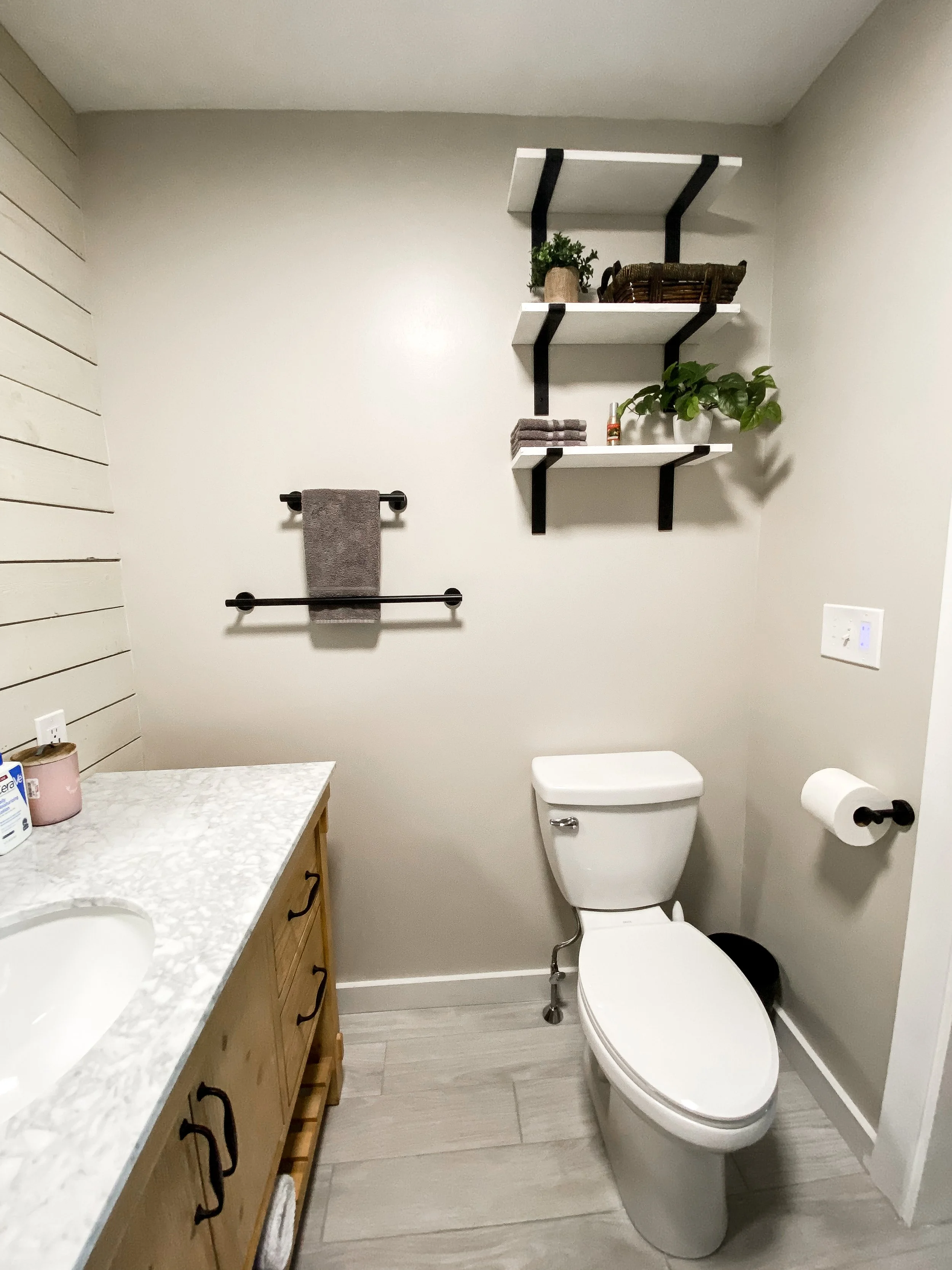 A small bathroom with a white toilet, a wooden vanity with a marble countertop, open shelves with plants and decor, and a gray towel hanging on a black towel bar.