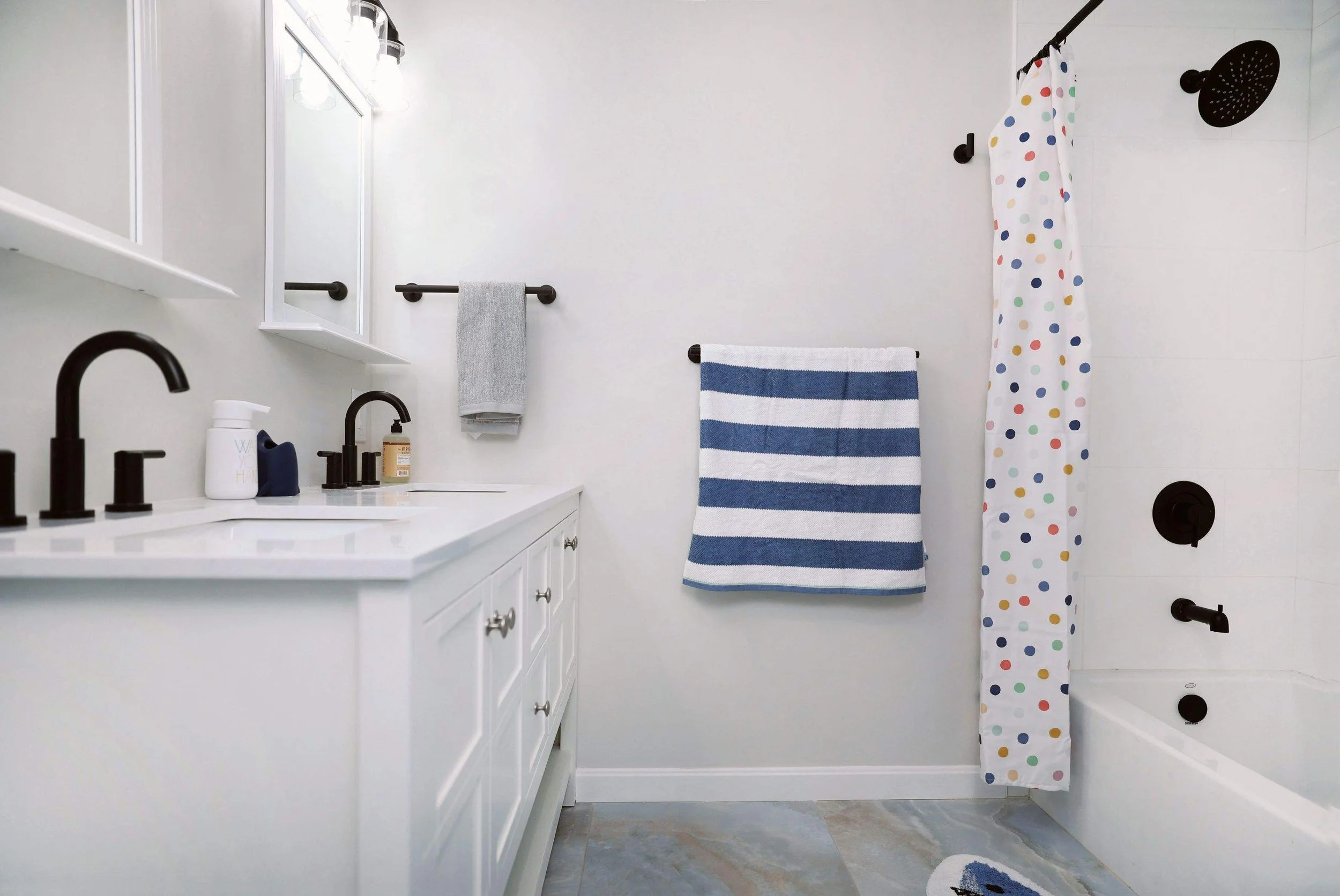 Bathroom with white vanity, black faucets, wall-mounted mirrors, gray and striped towels, polka-dot shower curtain, black showerhead, and white bathtub.