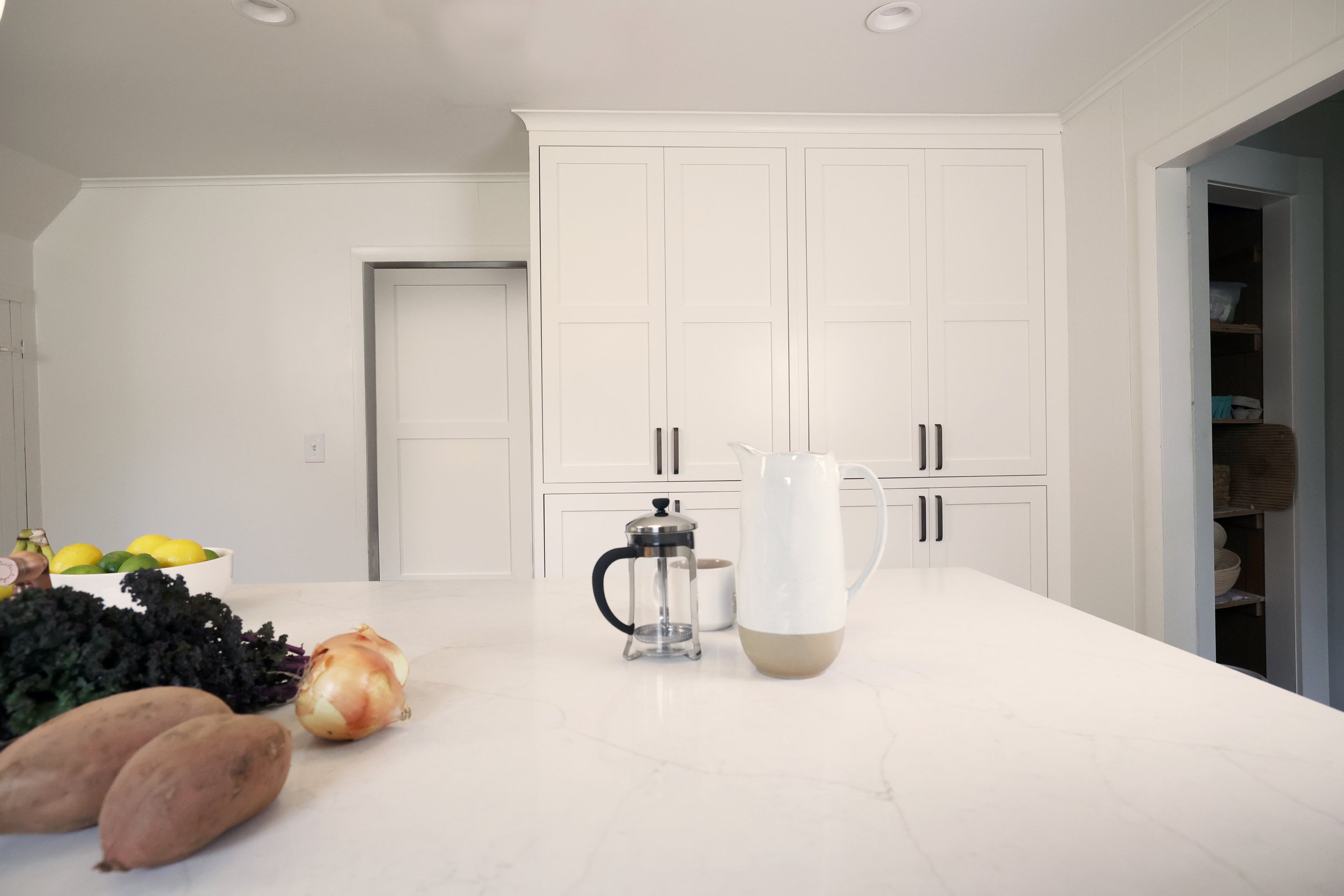 A white kitchen counter with a white pitcher, a French press, a mug, and some vegetables including sweet potatoes, onions, and kale. In the background, white cabinets and an open shelf with folded towels and bowls.