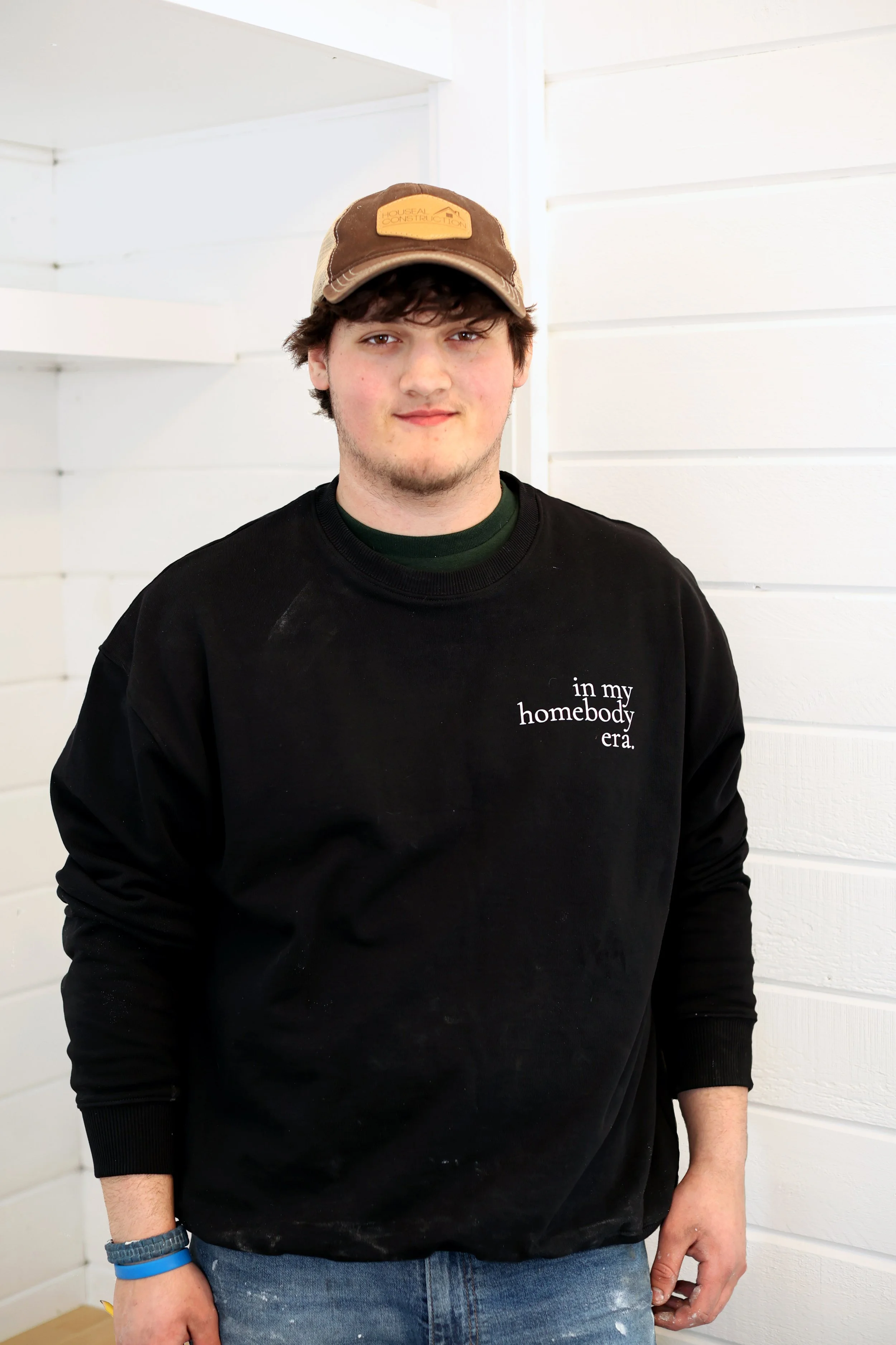 Young man wearing a brown and beige cap, black sweatshirt with white text, and jeans, standing against a white wooden wall.