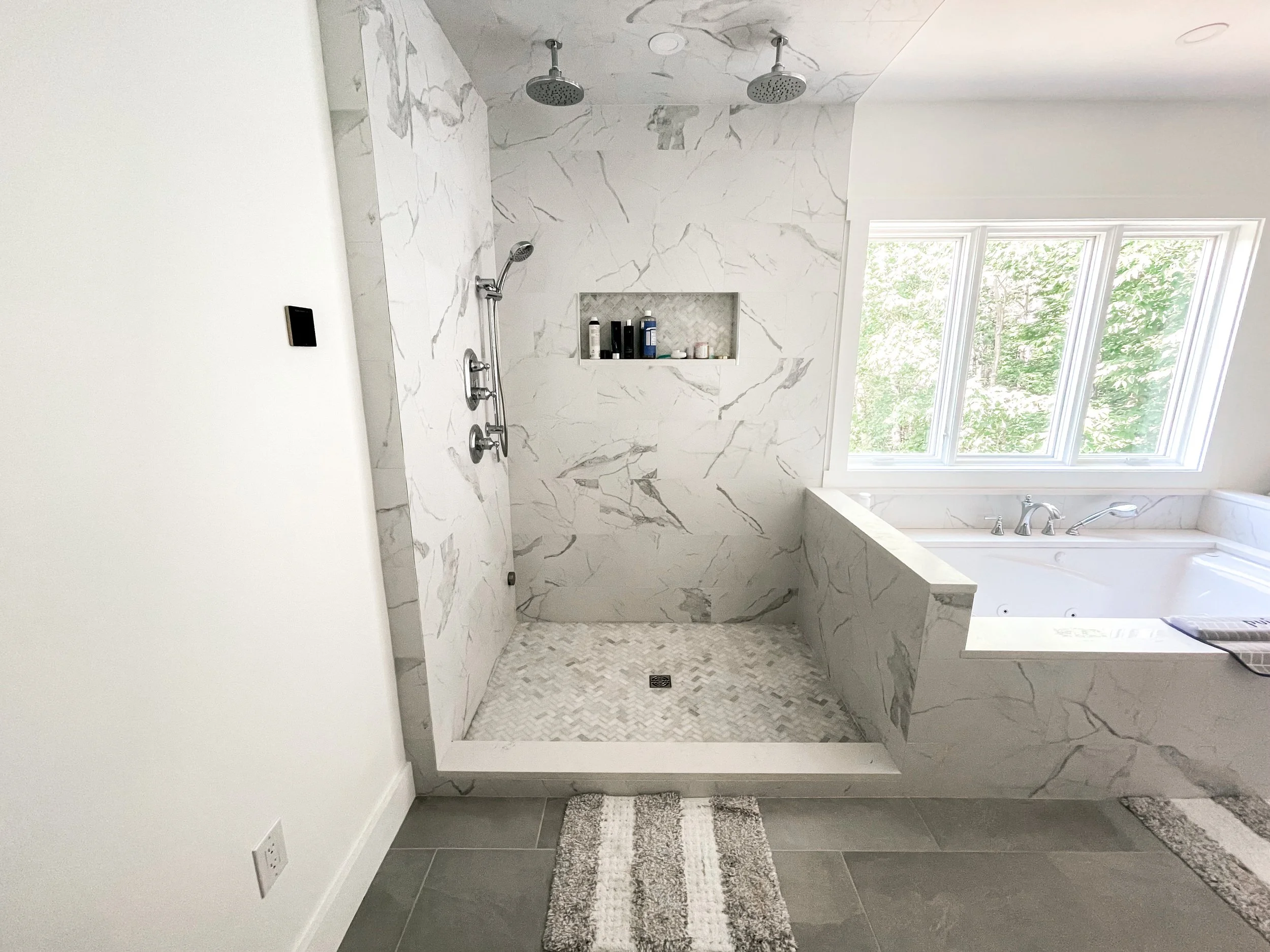 Bathroom with a walk-in shower featuring marble walls and a built-in niche, and a large bathtub beneath windows with a view of greenery outside.