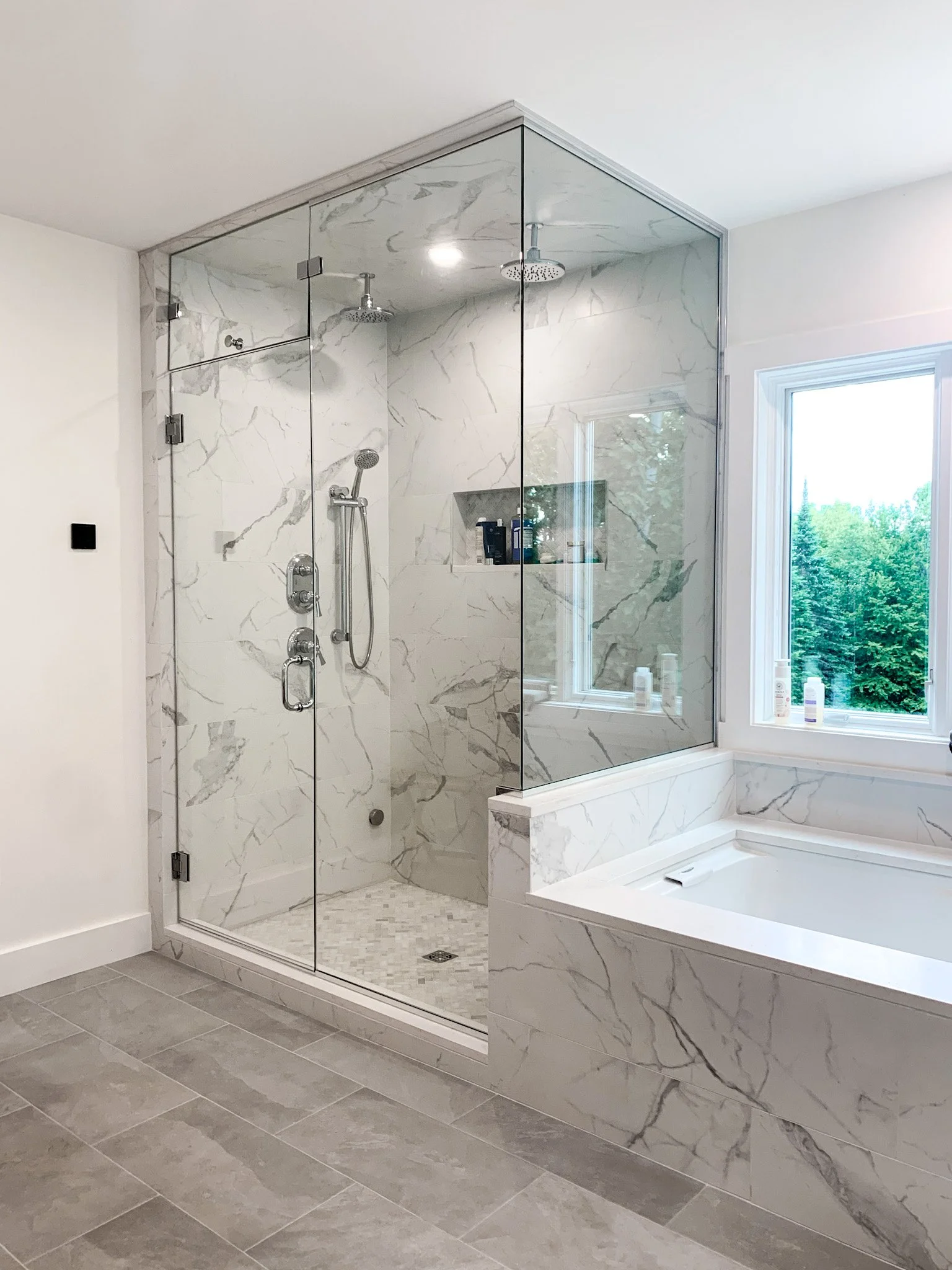 Modern bathroom with a glass-enclosed shower featuring marble walls and a separate bathtub near a window with greenery outside.
