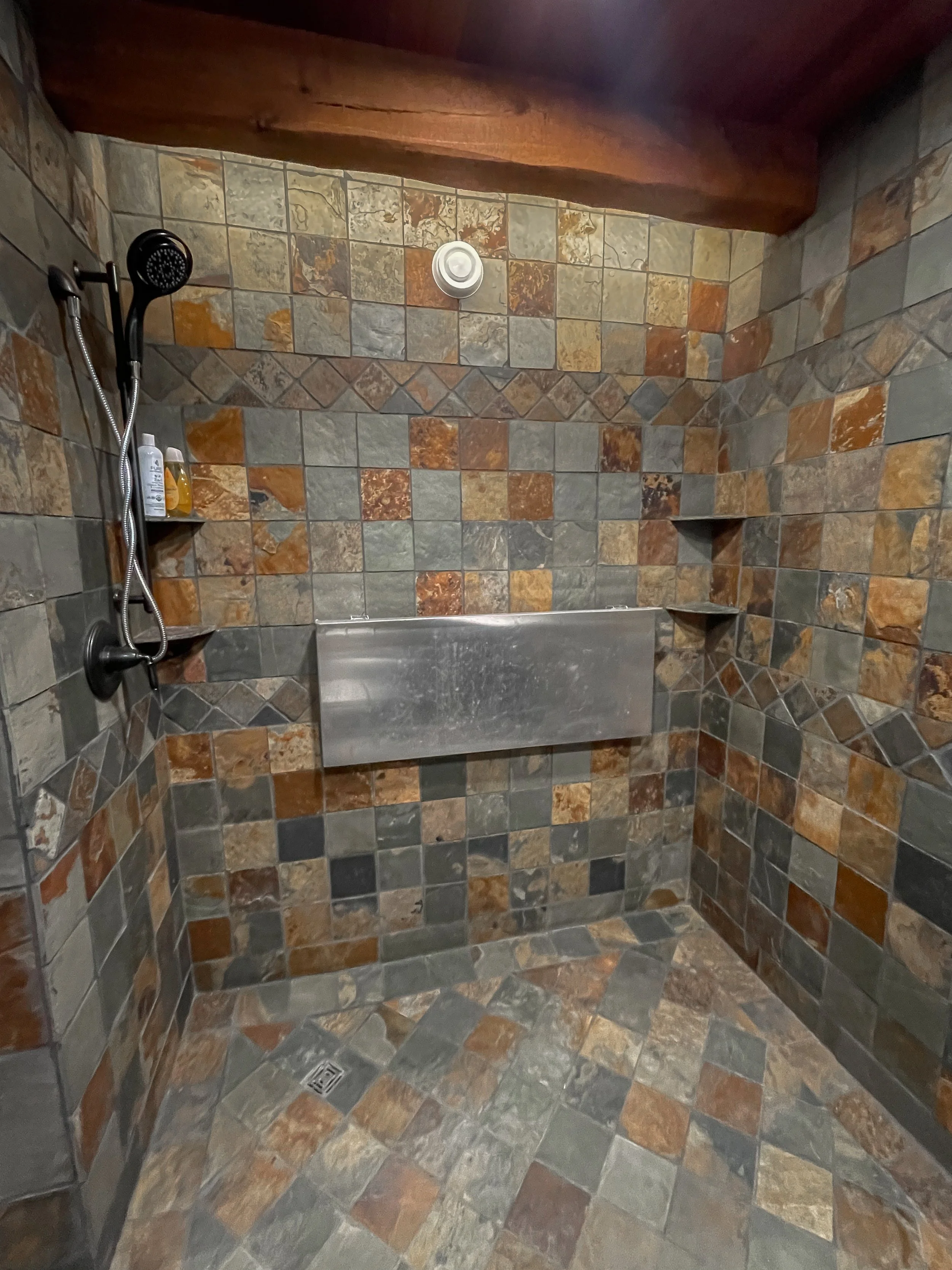 A walk-in shower with multicolored stone tile walls and floor, equipped with a black handheld showerhead, bottles of shampoo and soap on a small shelf, a metal shower bench, and a ceiling vent.