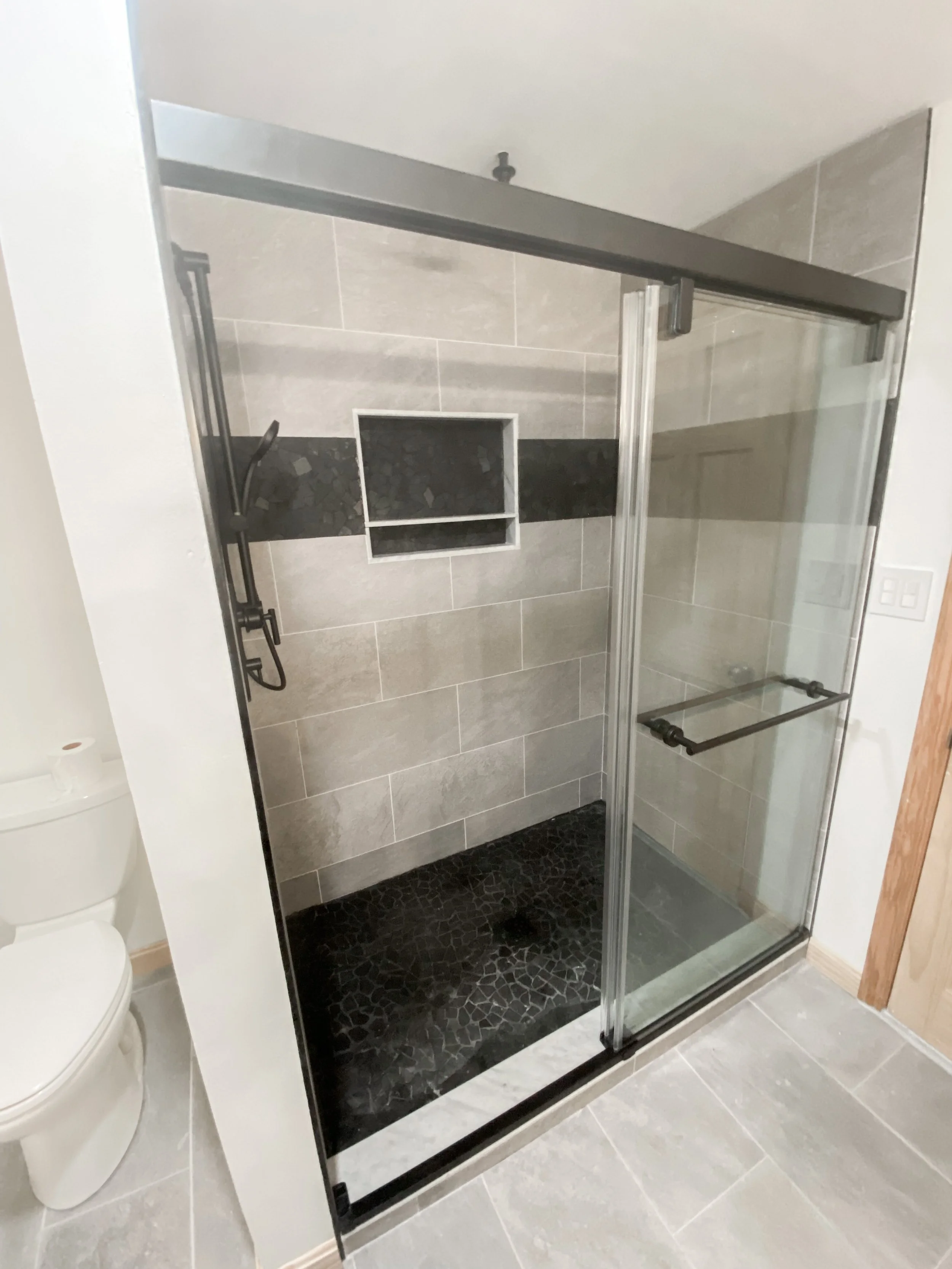 Shower with glass door, tiled walls, black pebble floor, and built-in shower niche in a bathroom.
