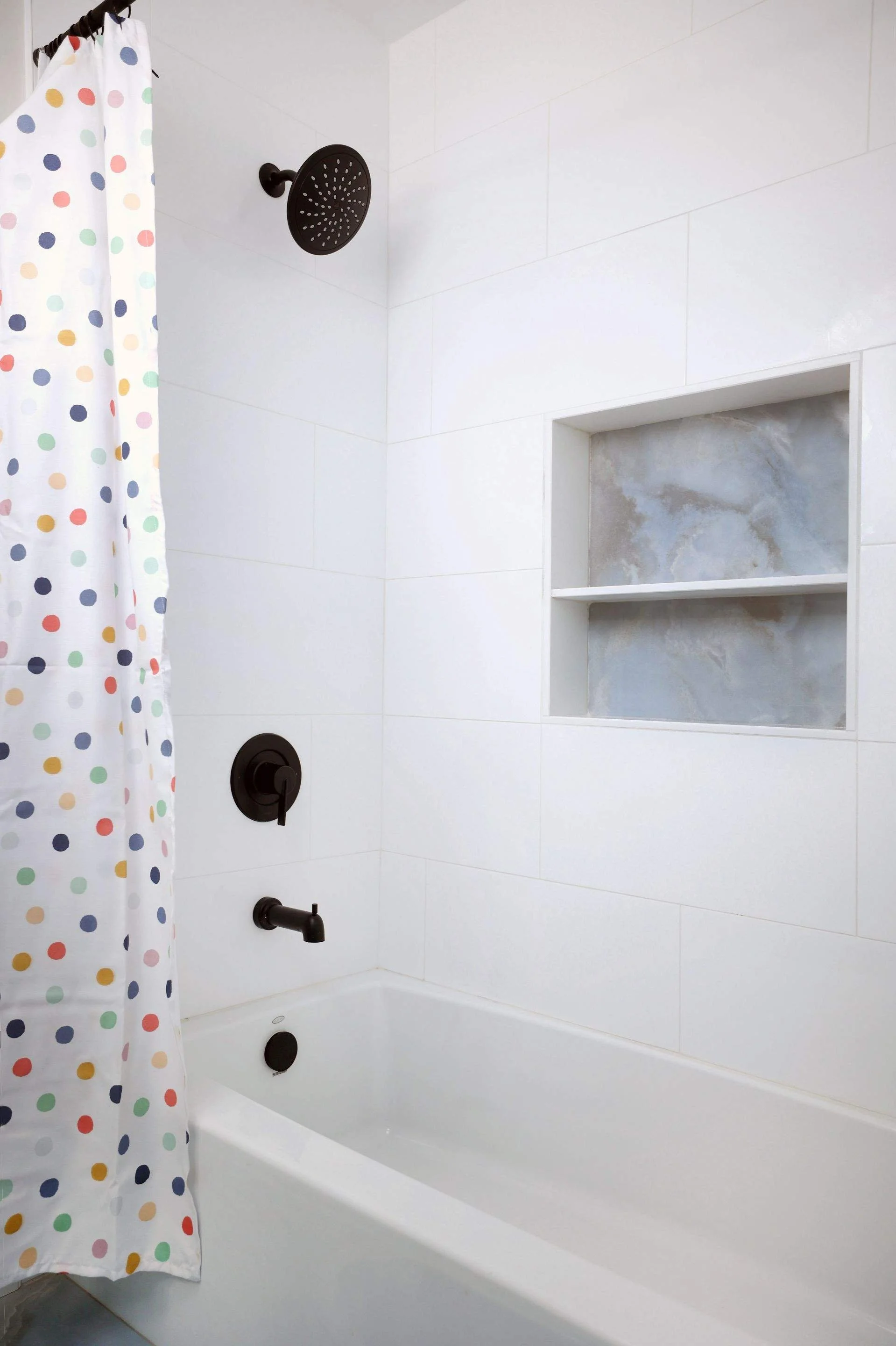 White bathtub with black showerhead, faucet, and controls, a colorful polka dot shower curtain, and a built-in tiled shelf in a bathroom with white tiled walls.