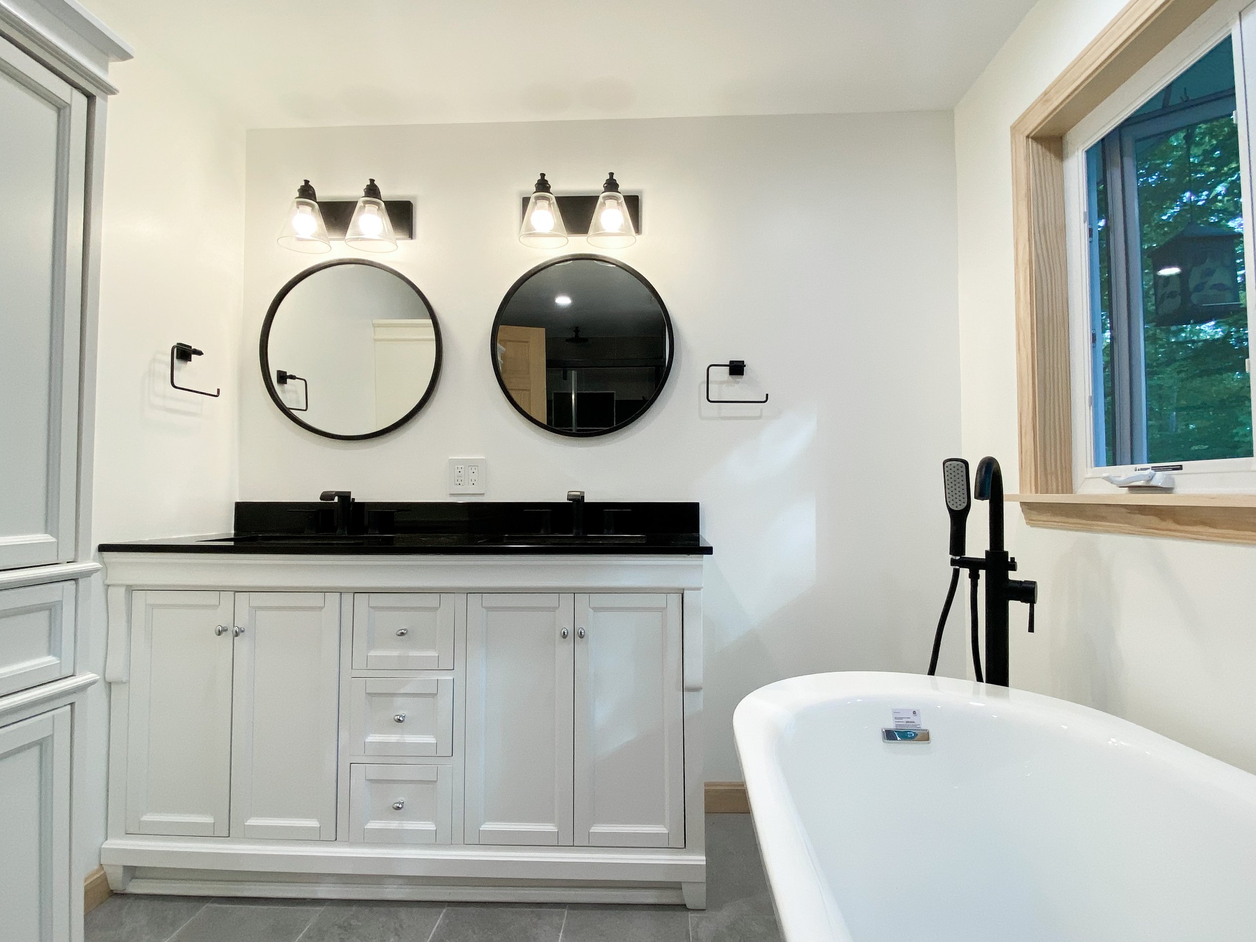 Modern bathroom with dual mirrors, black double sink vanity, black faucet, white cabinetry, bathtub, window showing green outdoor trees, and wooden window trim.