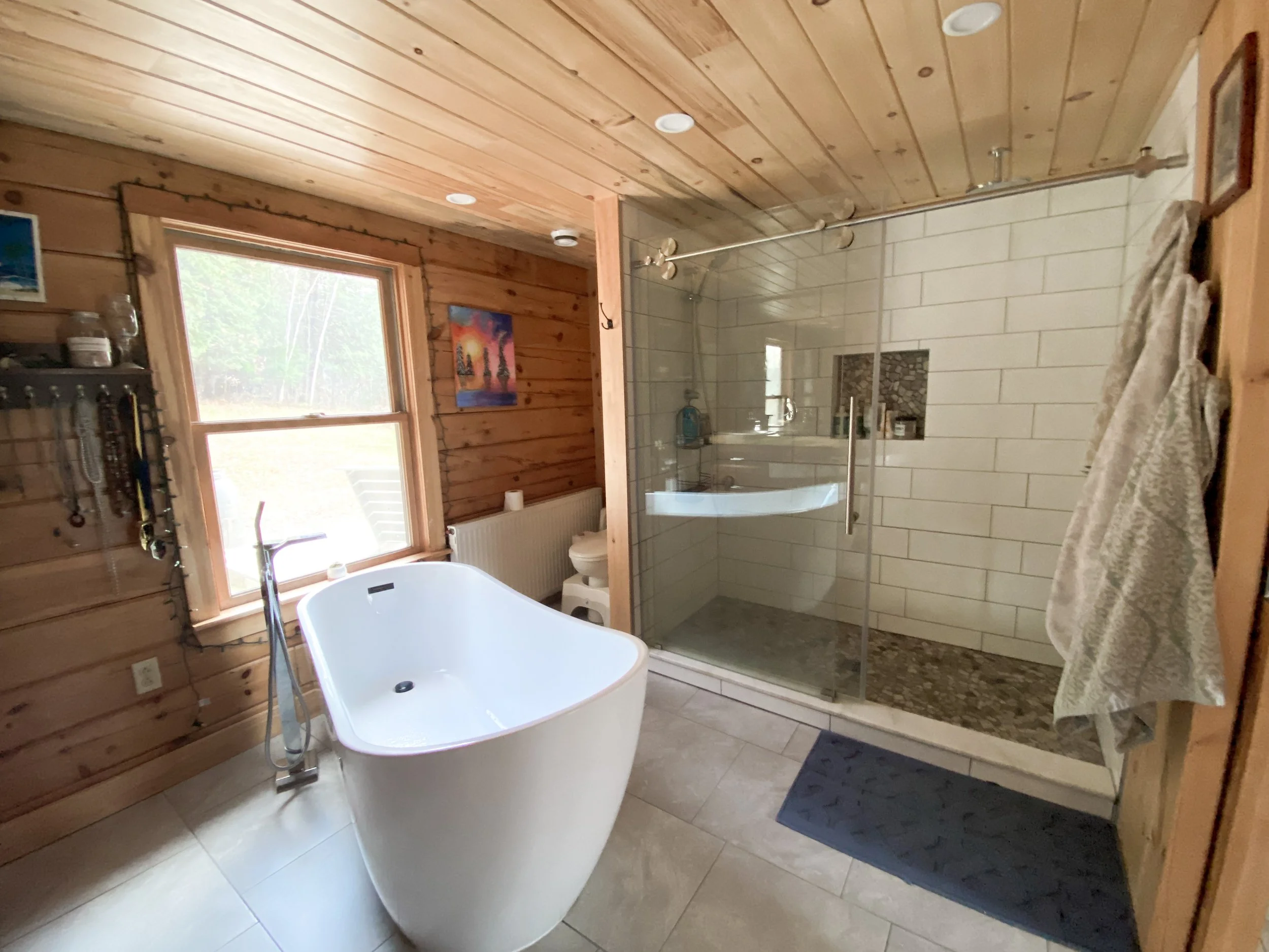 Modern bathroom with a freestanding bathtub, shower with glass door, wooden wall panels, and a window showing trees outside.