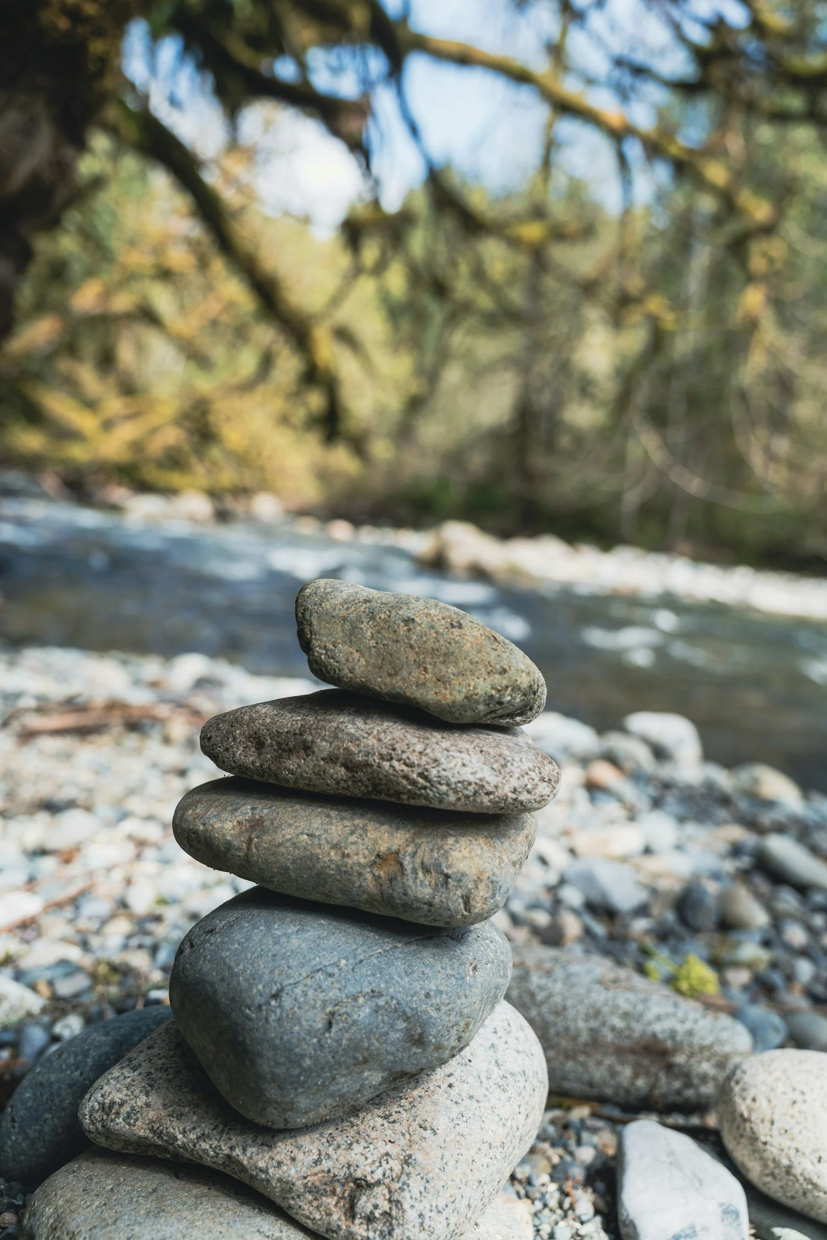 Pyramid of smooth stones stacked on a rocky riverbank, with a blurred background of trees and water.