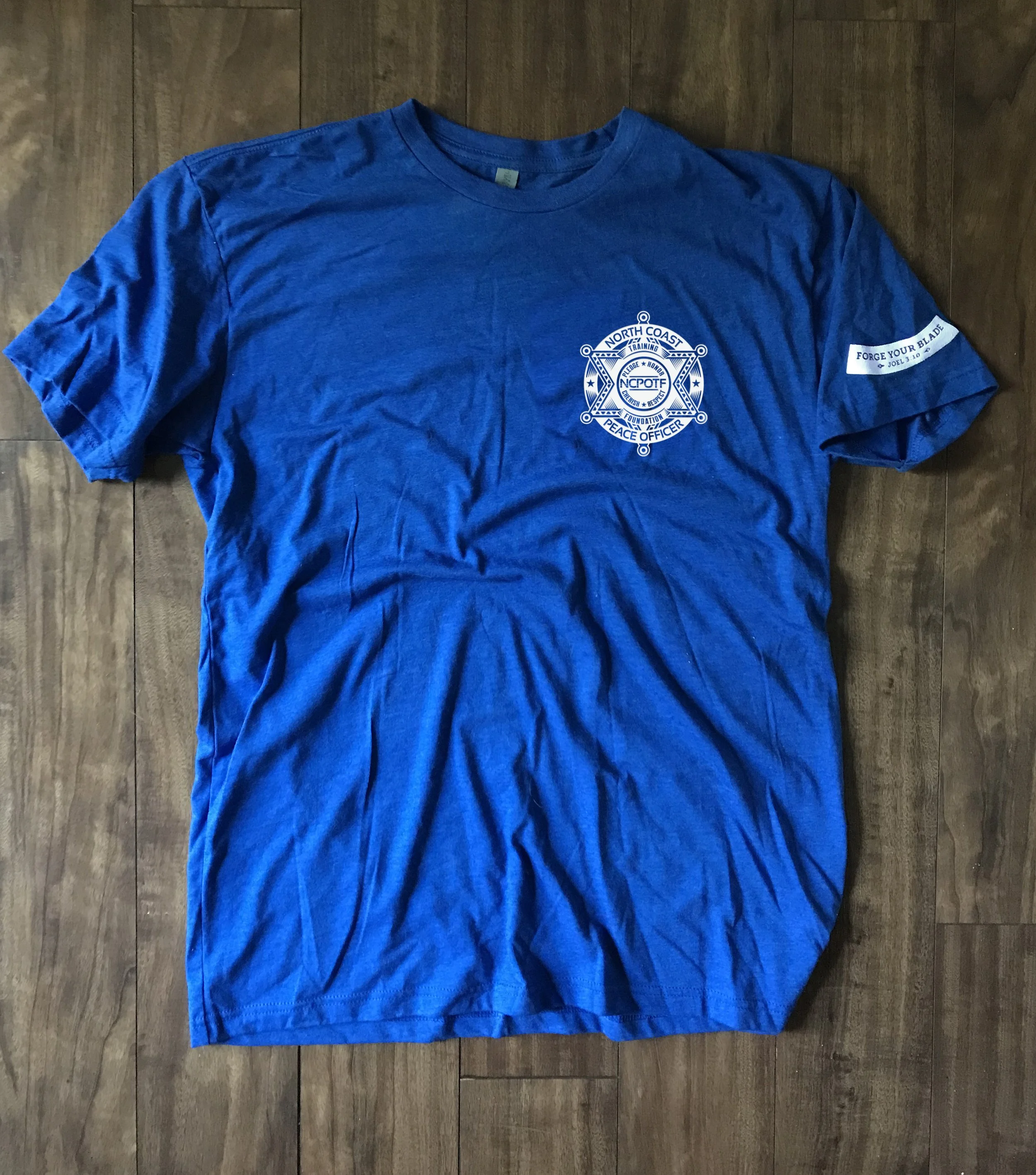 Blue T-shirt with North Coast Peace Officer badge logo on the front left chest and a patch on the right sleeve reading 'Forge Your Blade' with Ephesians 6:17