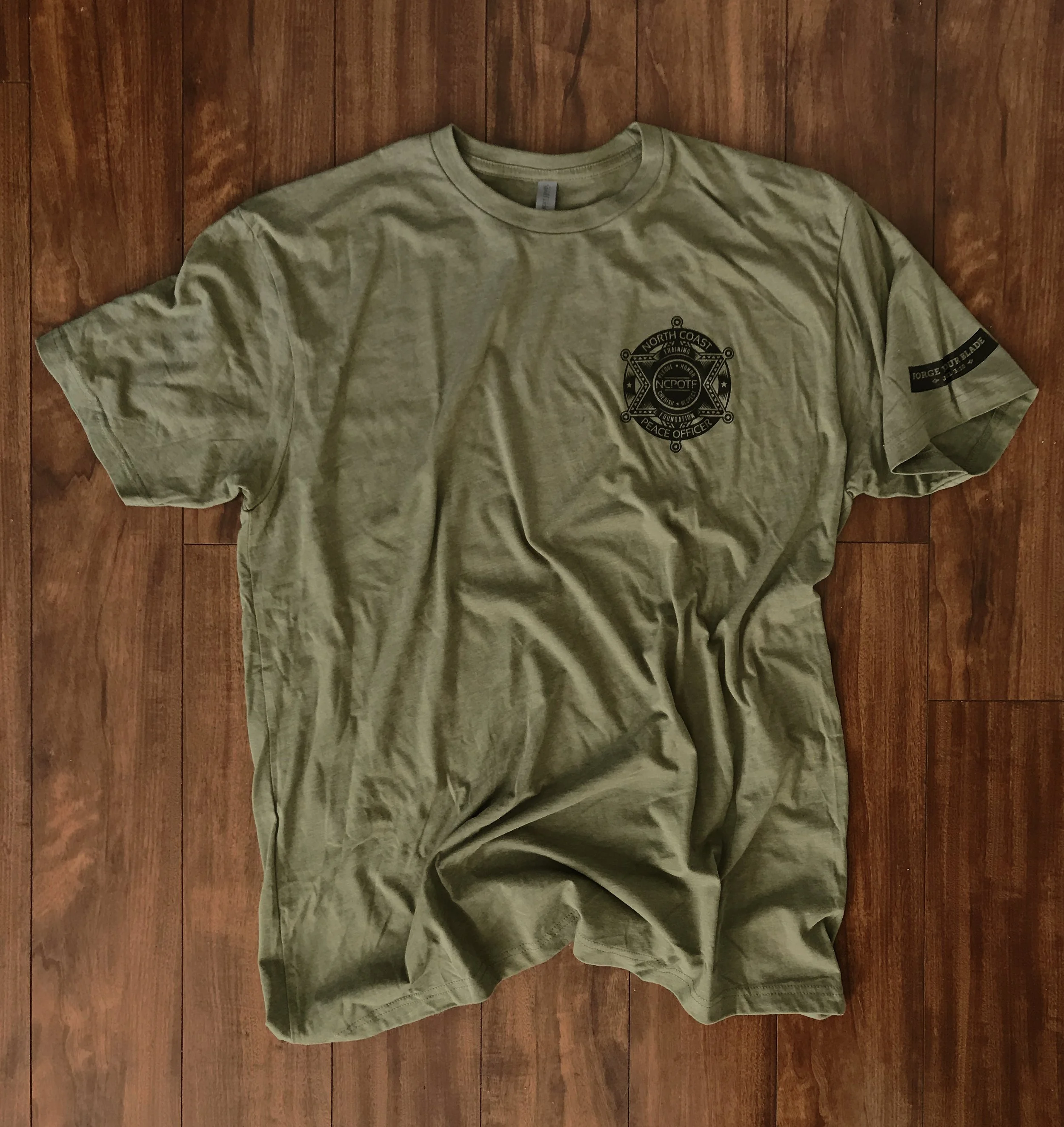 A beige t-shirt with a black badge-like emblem on the chest and a black patch on the sleeve, laid flat on a wooden floor.