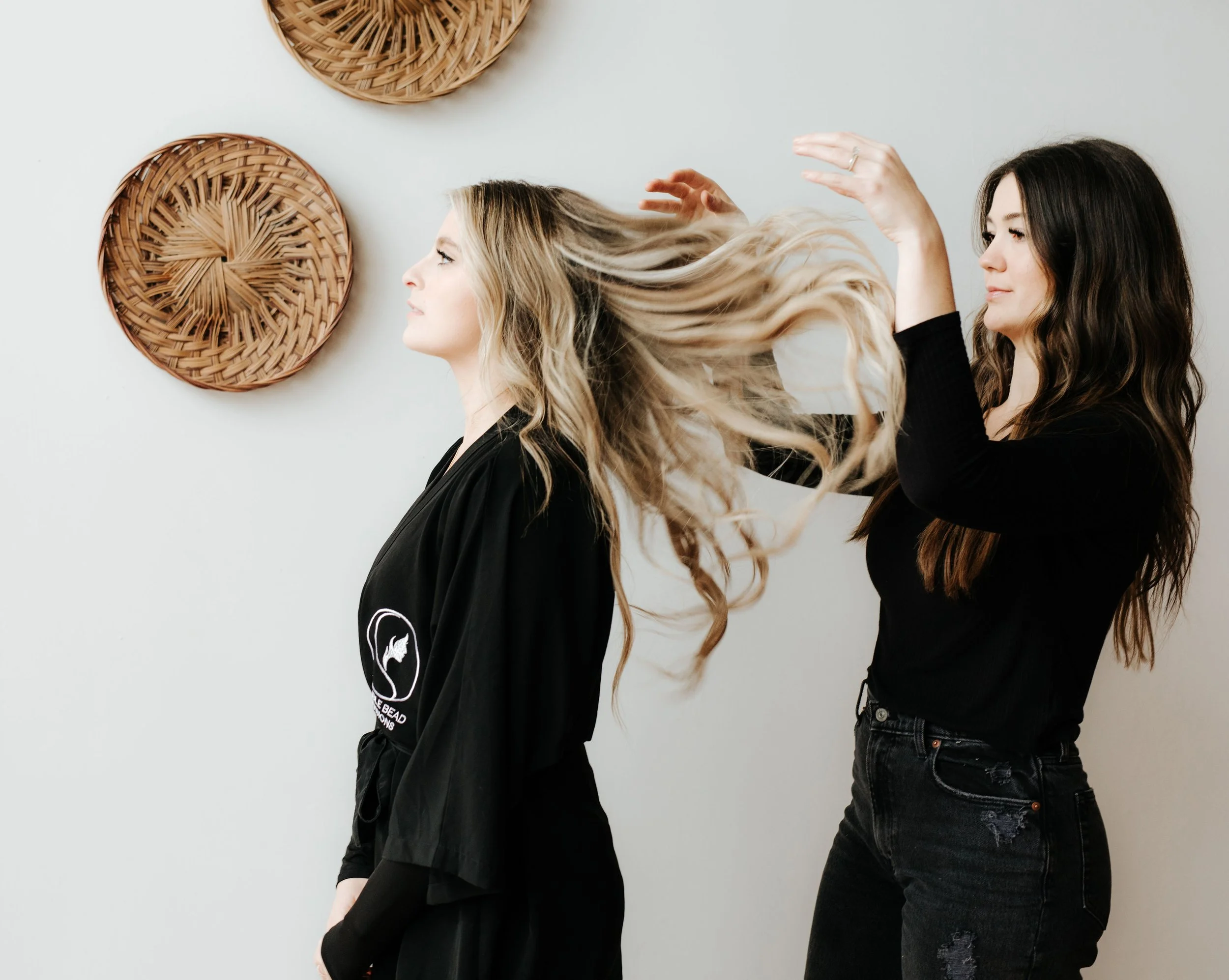 A stylist brushing the long blonde hair of a woman with dark blonde hair against a white wall decorated with woven wall hangings.