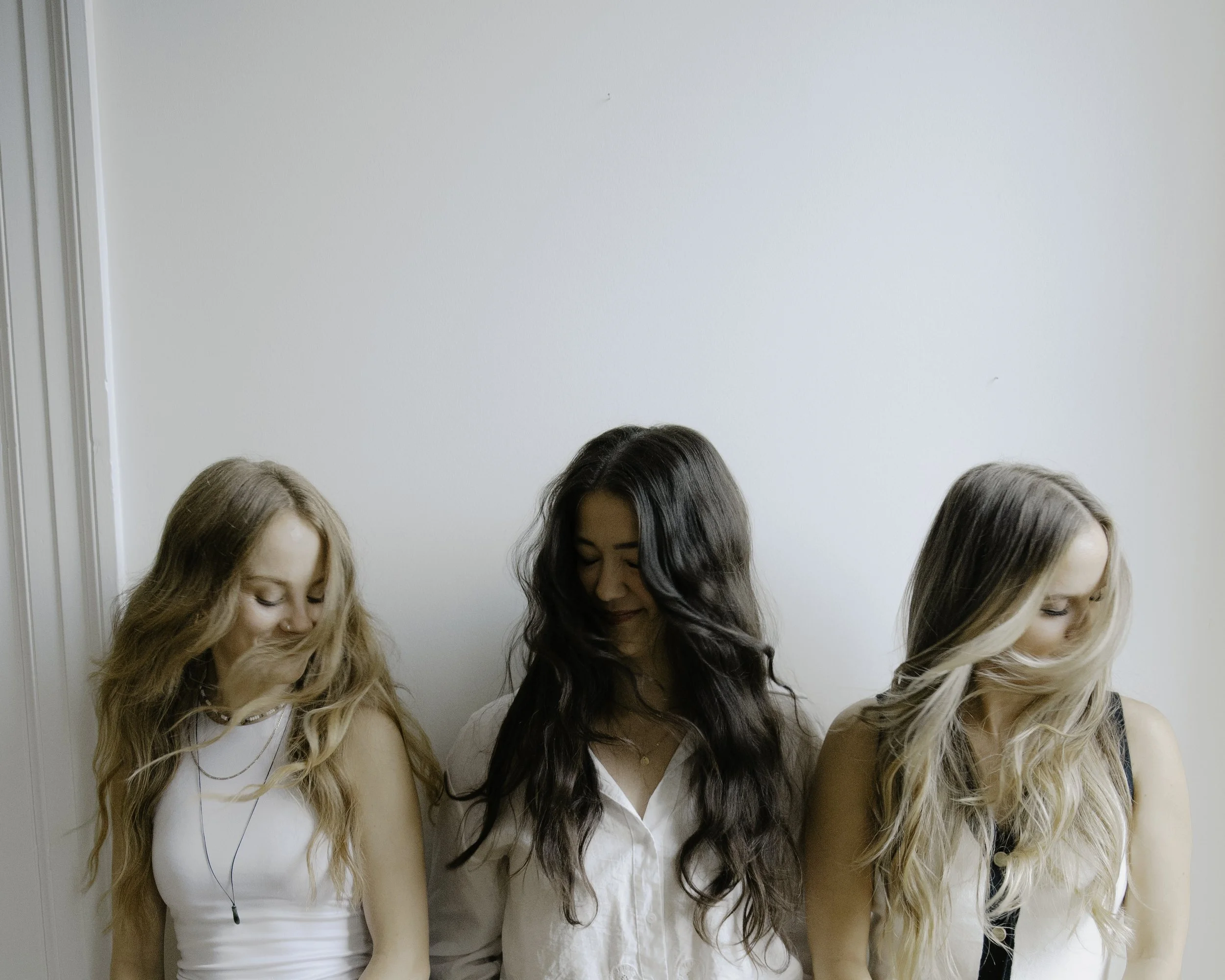 Three young women standing together, each with long hair, in front of a plain white wall.