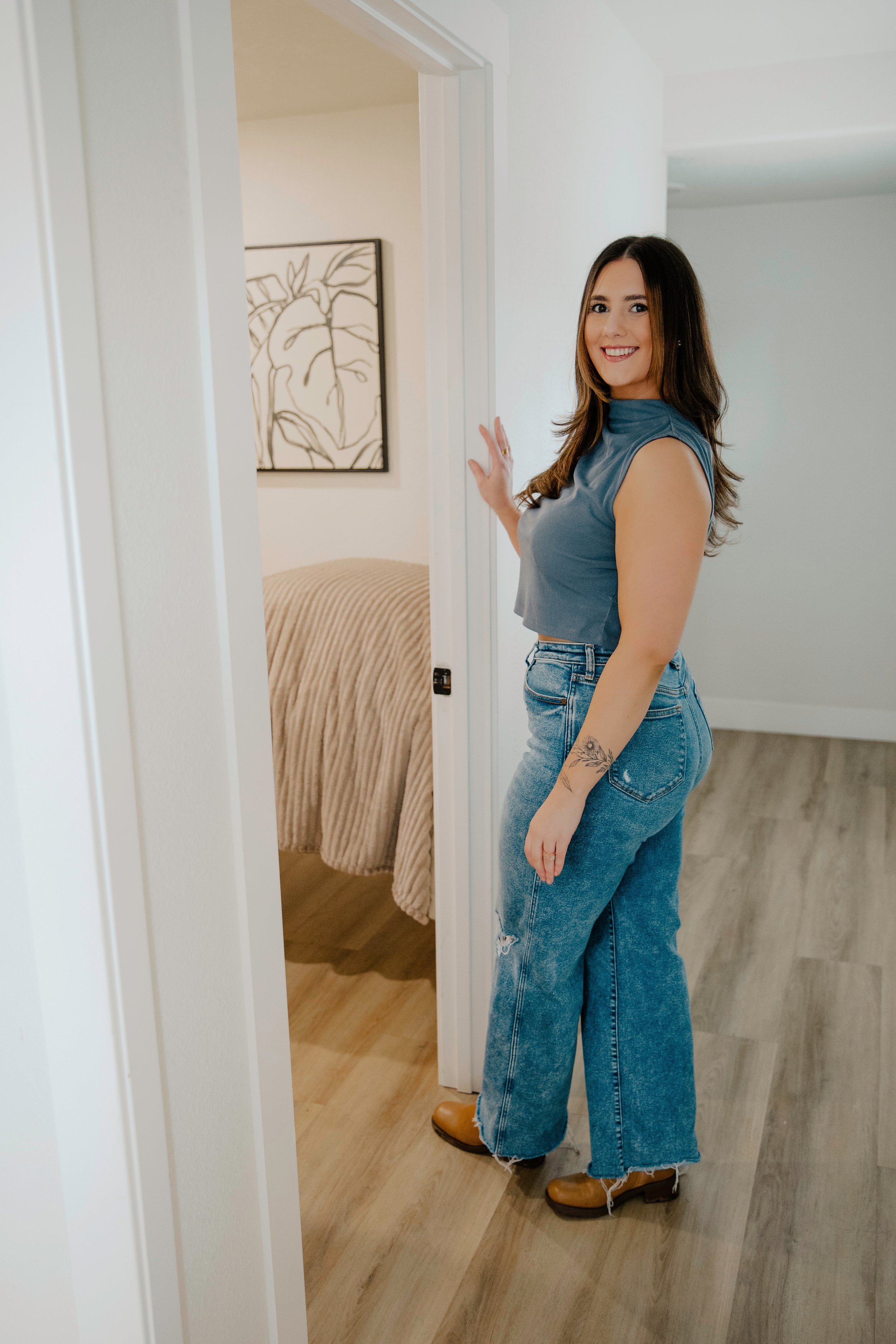 A woman with long brown hair, wearing a sleeveless blue top, ripped wide-leg jeans, and brown boots, standing in a doorway and smiling at the camera inside a home.