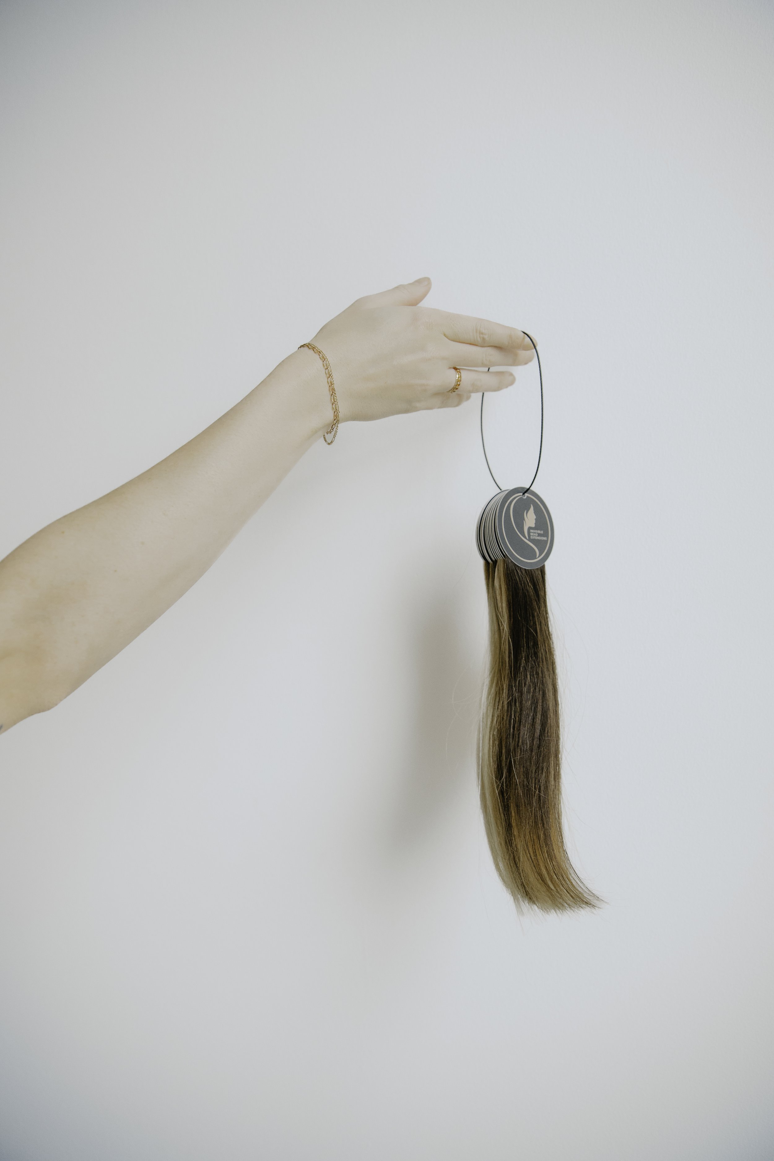 A hand holding a hair color sample with hair attached, against a plain white wall.