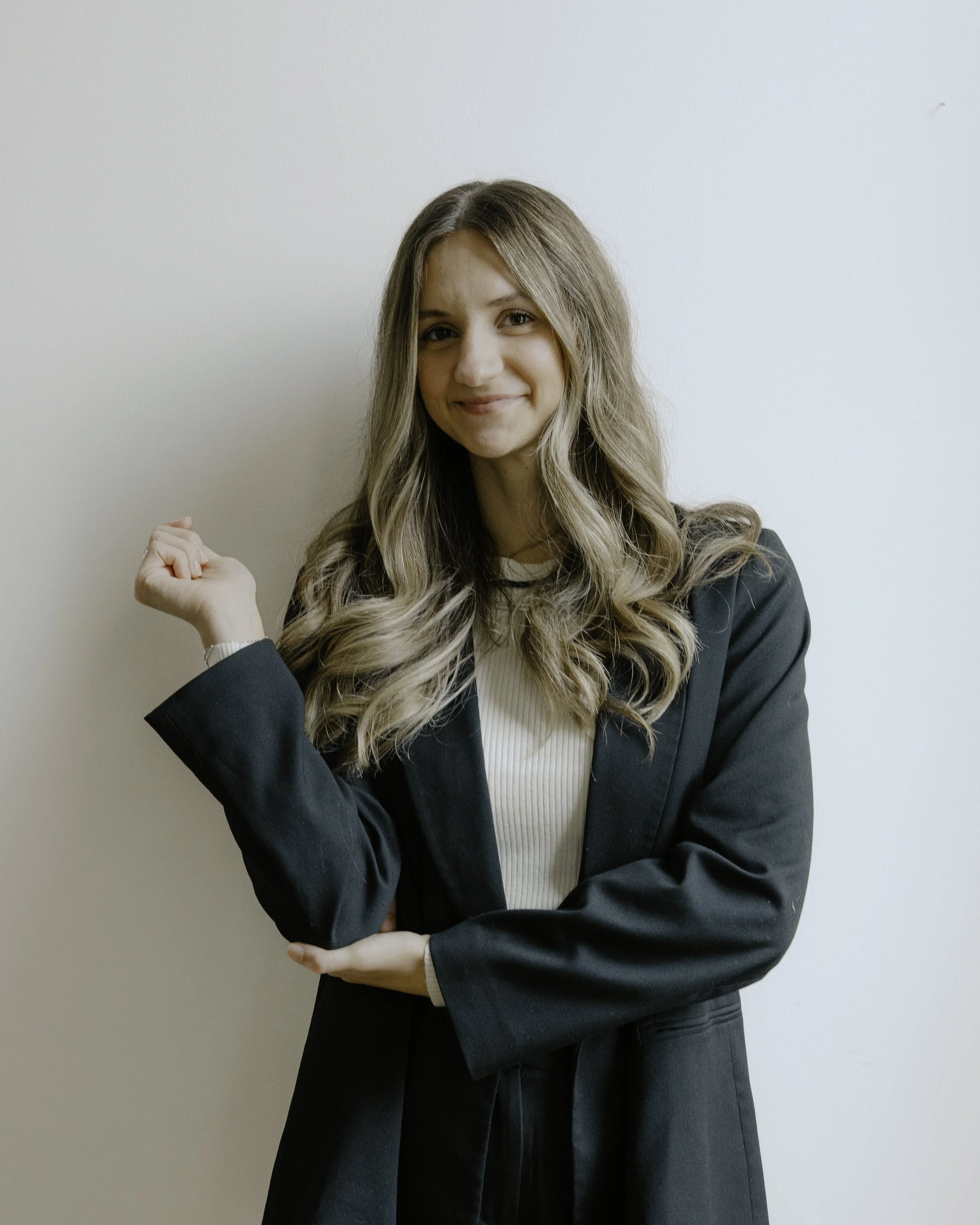 A young woman with long wavy blonde hair, wearing a black blazer over a white top, standing against a plain white wall. She is smiling and making a fist with her right hand, celebrating a victory or success.