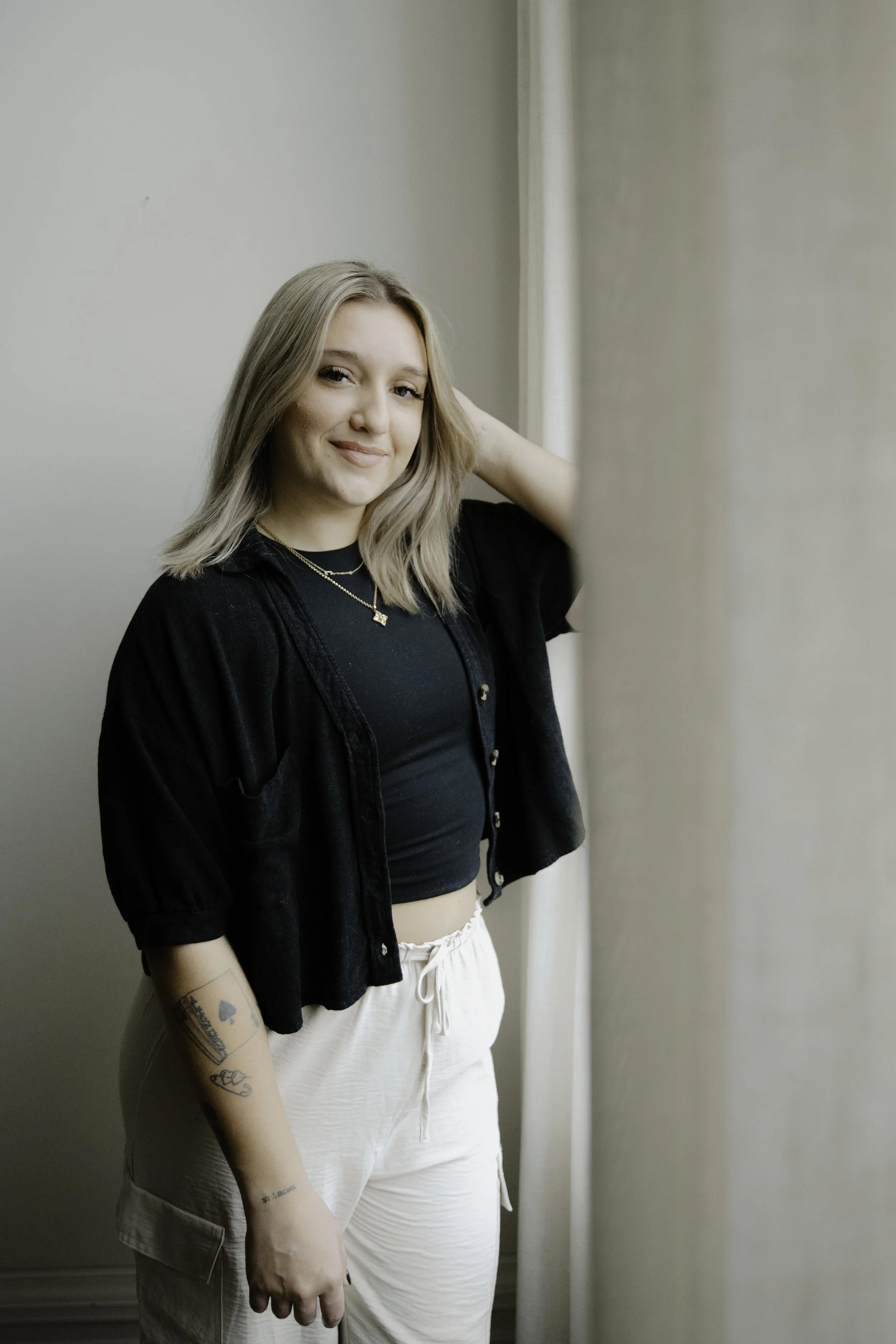 A young woman with shoulder-length blonde hair, wearing a black crop top, black button-up shirt, and beige pants, standing near a light-colored wall and window, smiling at the camera.