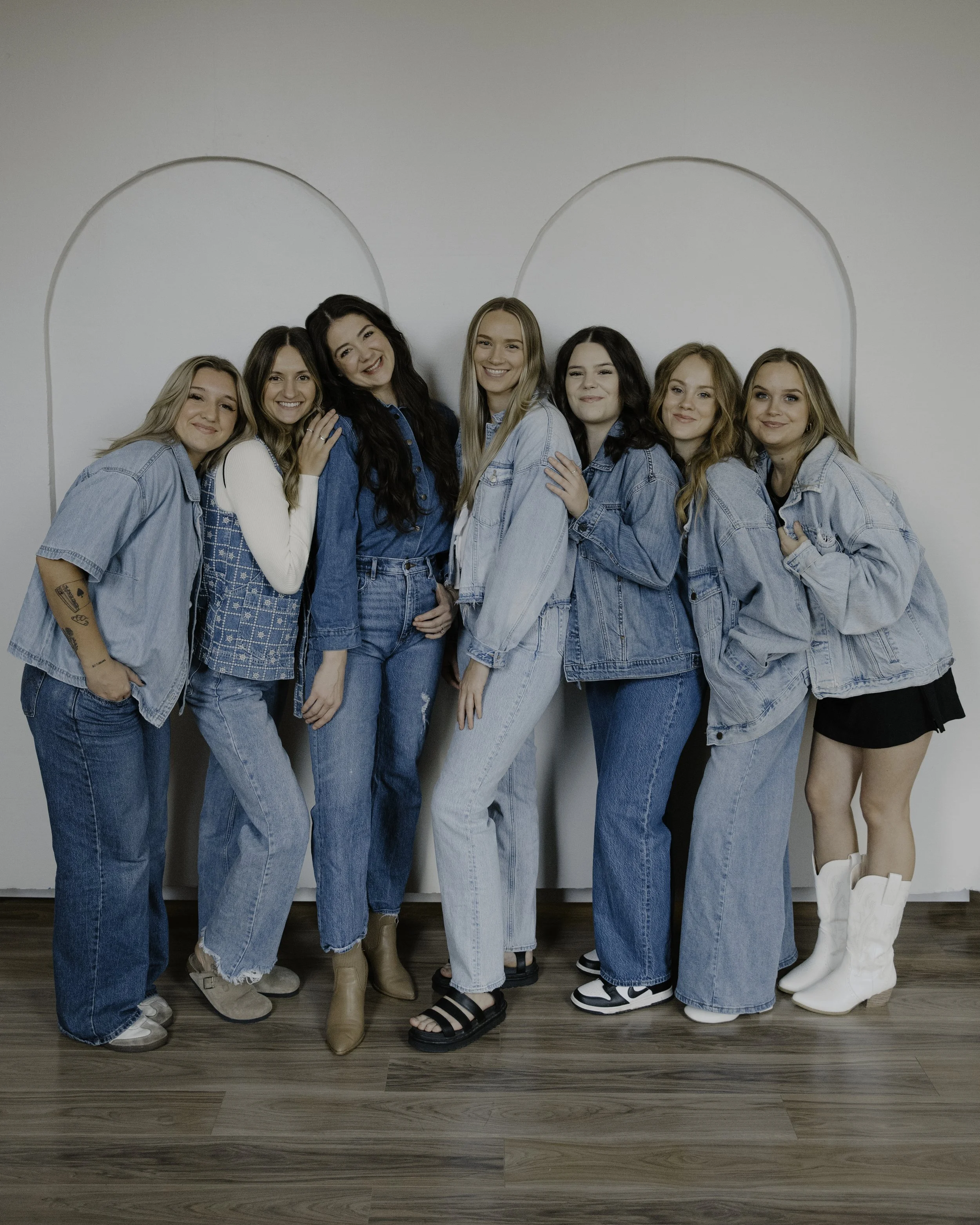 Group of seven women in casual denim and other styles of clothing, standing close together in front of a plain wall.