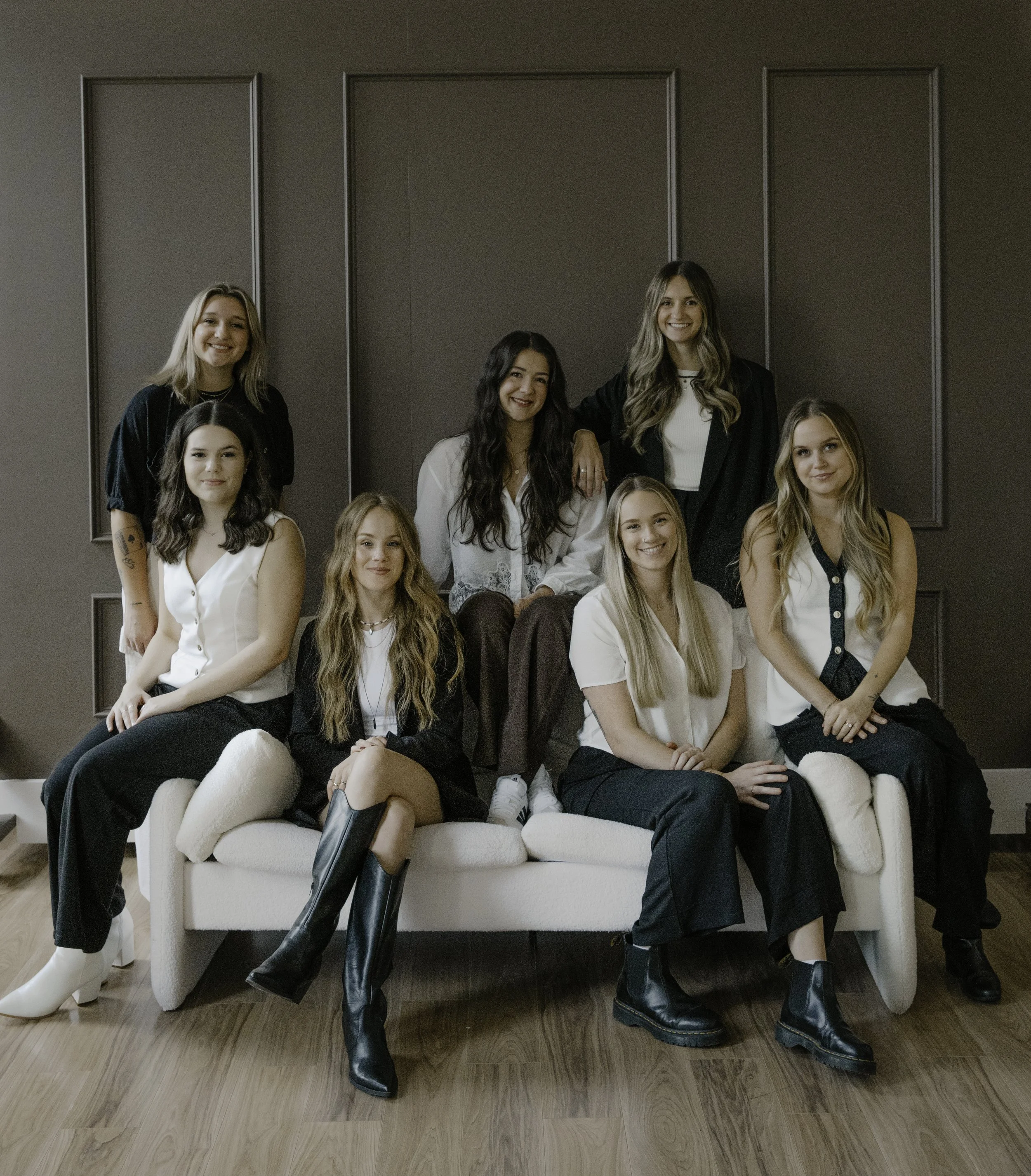 Group of eight women sitting and standing in a living room, smiling at the camera, dressed in casual and semi-formal clothing.