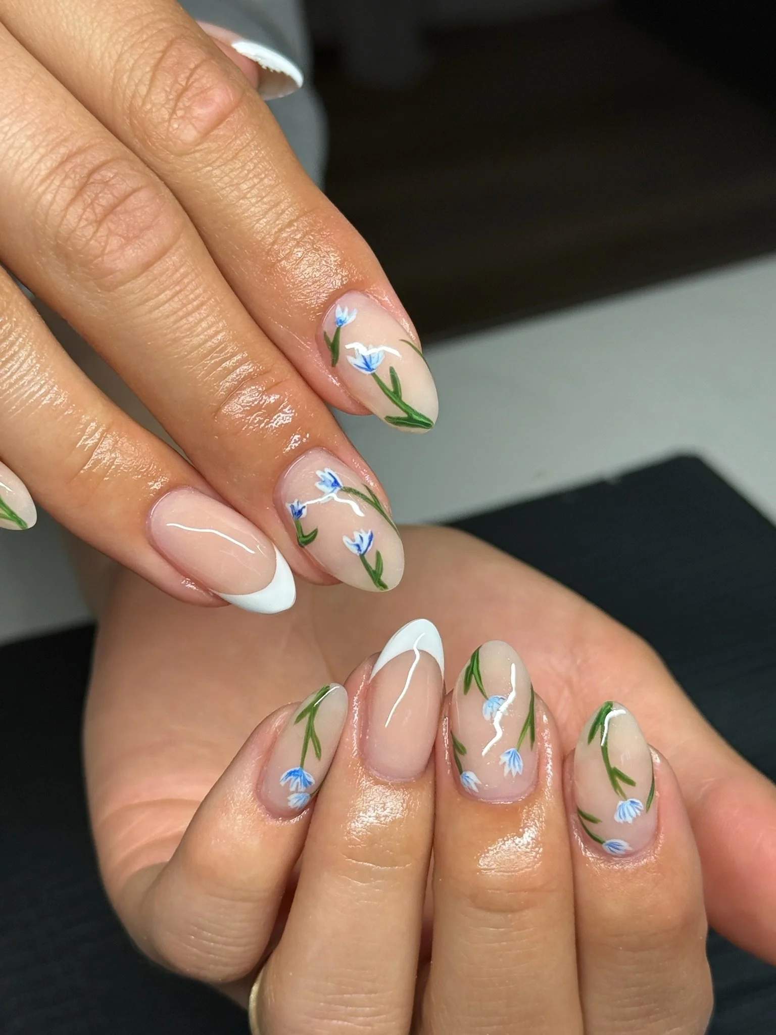 Nail art with a sheer nude base, white French tips, and hand-painted blue flowers with green stems on each nail.