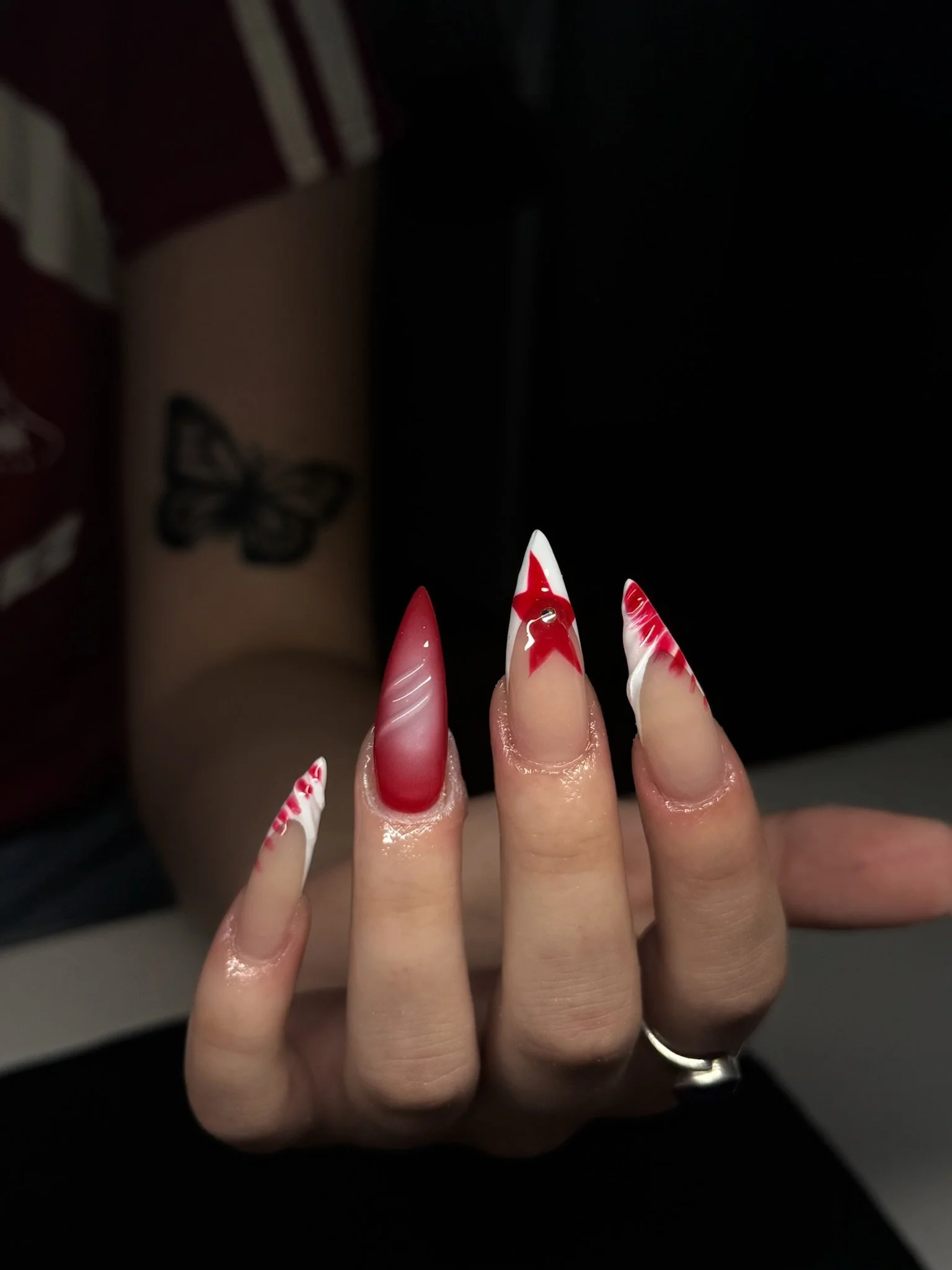 Close-up of a hand with long, decorated nails featuring red, white, and pink designs, with a butterfly tattoo on the arm in the background.