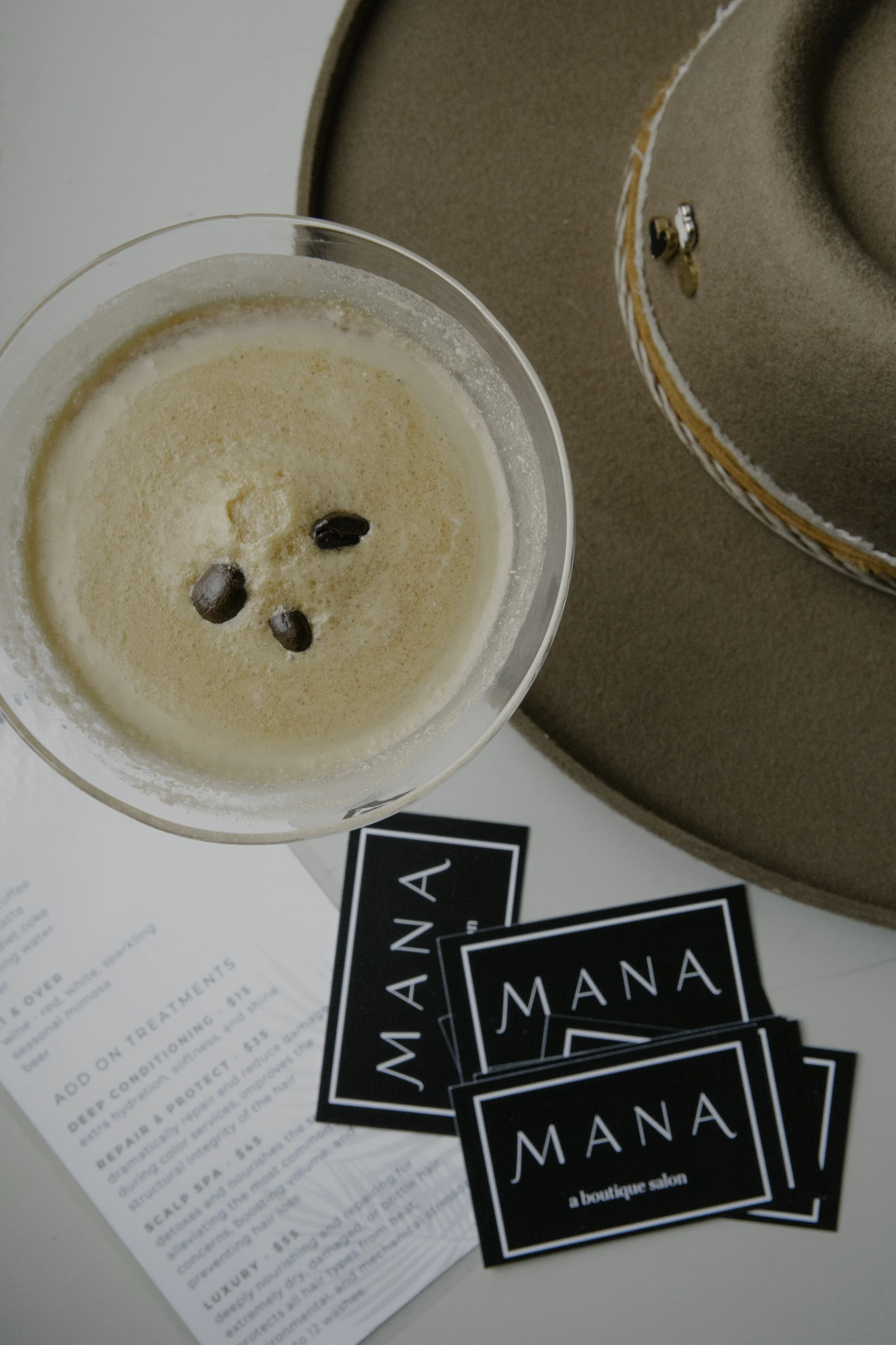 A glass of coffee with coffee beans on top, placed on a table next to business cards that say 'MANA, a boutique salon', with a brown hat in the background.