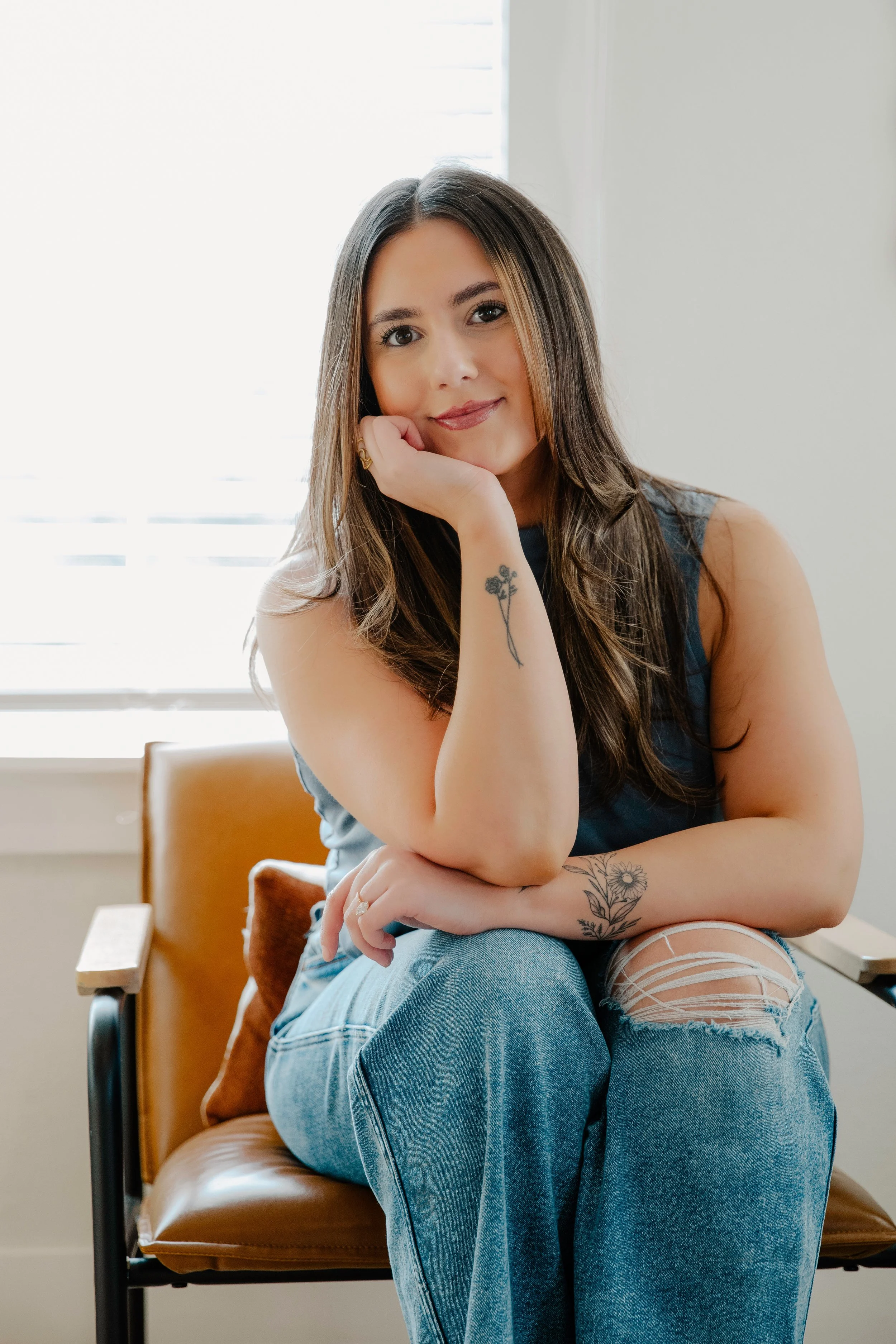 A young woman with long brown hair, tattoos on her arms, wearing a sleeveless top and ripped jeans, sitting on a chair near a window and smiling at the camera.