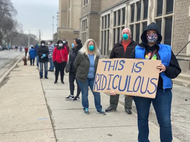 Milwaukee voters in long lines with masks  2020. Woman at front of line holding This Is Ridiculous" handmade sign. of line. Photo credit from the Milwaukee Journal Sentinal.