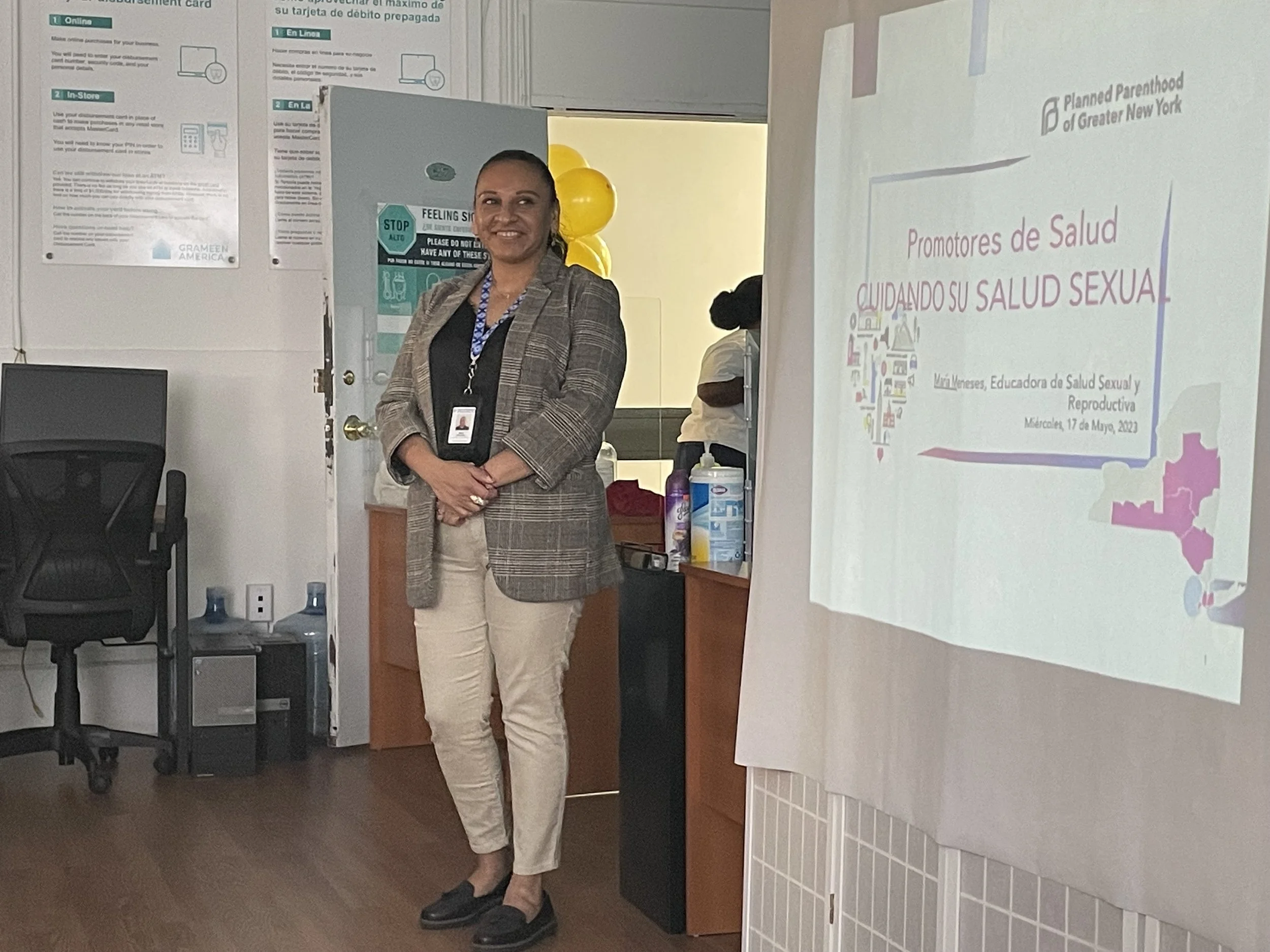 A person standing in an office next to a presentation screen. The screen features a slide from Planned Parenthood of Greater New York titled 'Promotores de Salud CUIDANDO SU SALUD SEXUAL.' The office contains office furniture and some posters on the wall.