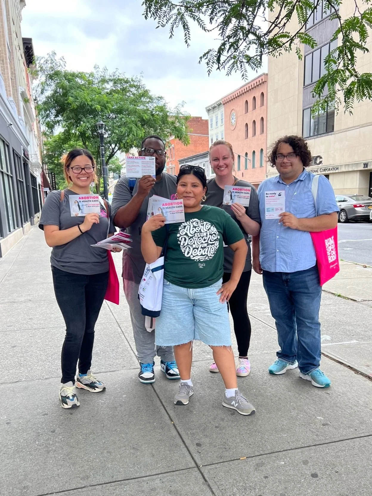 Group of five people standing on a city sidewalk, holding flyers that read "Abortion is Health Care," with buildings and trees in the background.