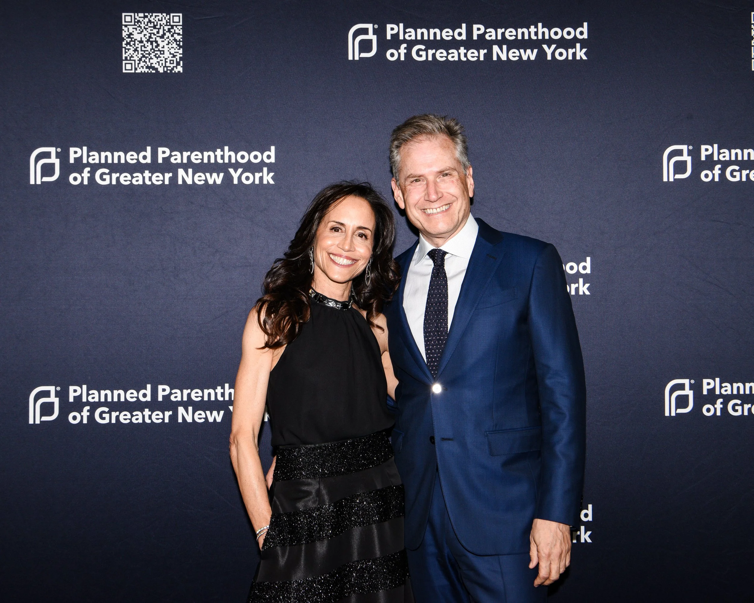 A man and a woman posing together at a Planned Parenthood of Greater New York event, standing in front of a backdrop with logos and a QR code.