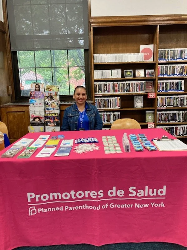 A woman sitting at a table covered with a pink tablecloth that says 'Promotores de Salud, Planned Parenthood of Greater New York.' The table has brochures, pamphlets, pens, and other informational materials. Behind her is a shelf with DVDs and a magazine rack.