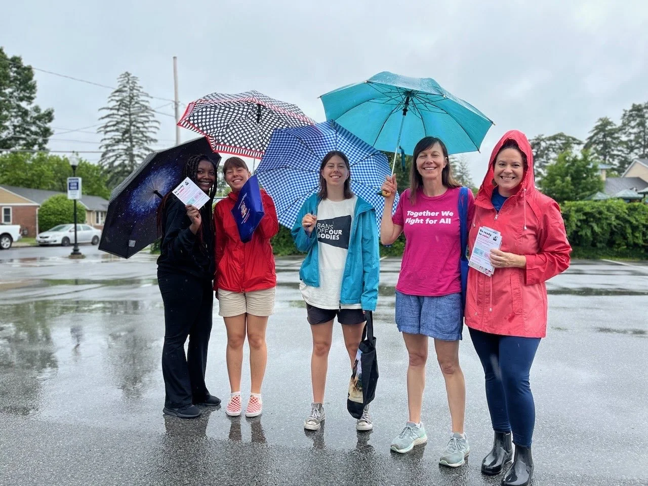 Group of five women standing together outdoors on a rainy day, each holding an umbrella and smiling, wearing casual and rain attire.