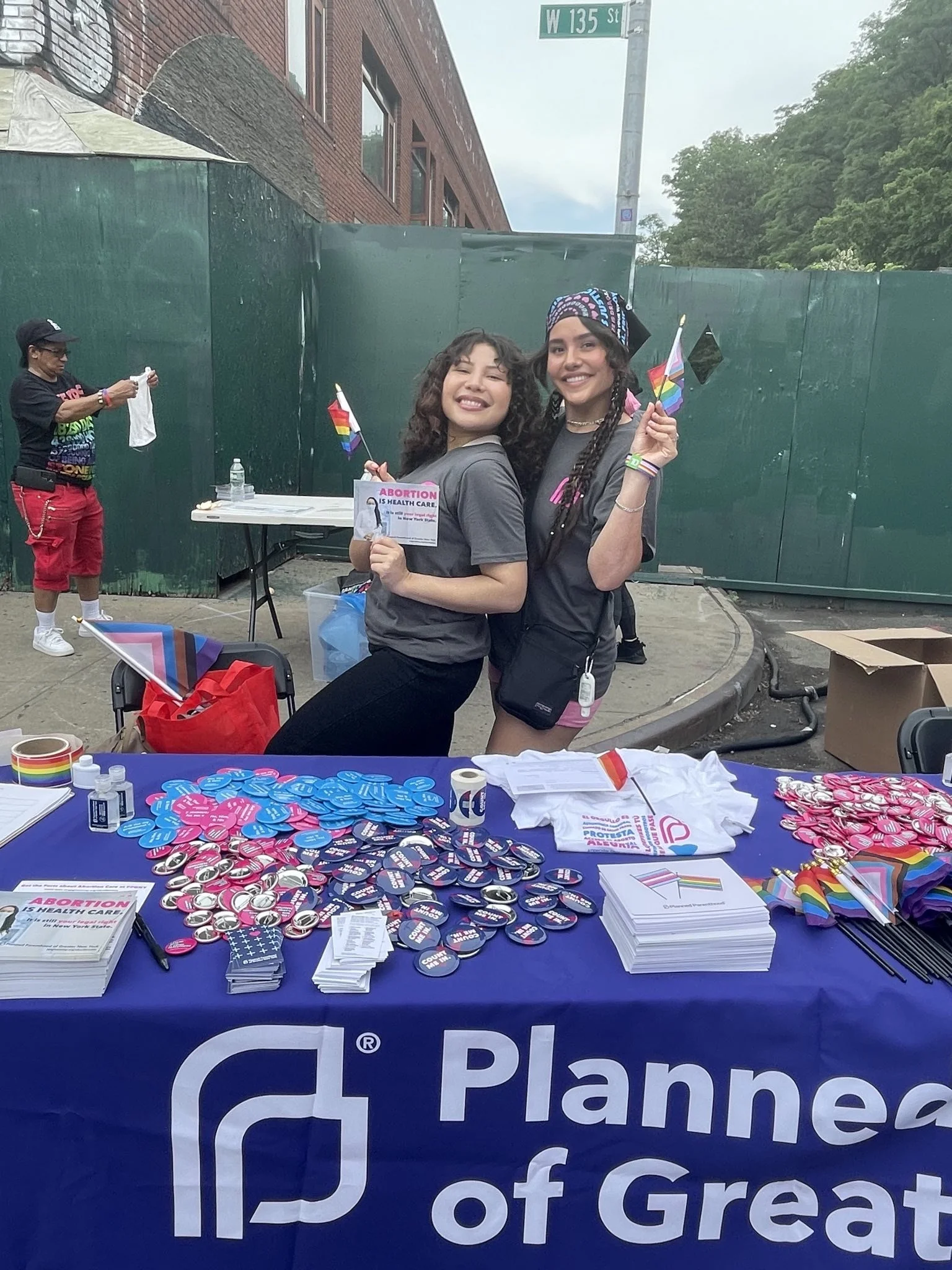 Two people smiling and holding rainbow flags at a Planned Parenthood table with informational materials, buttons, and shirts on display. The table is located outdoors.