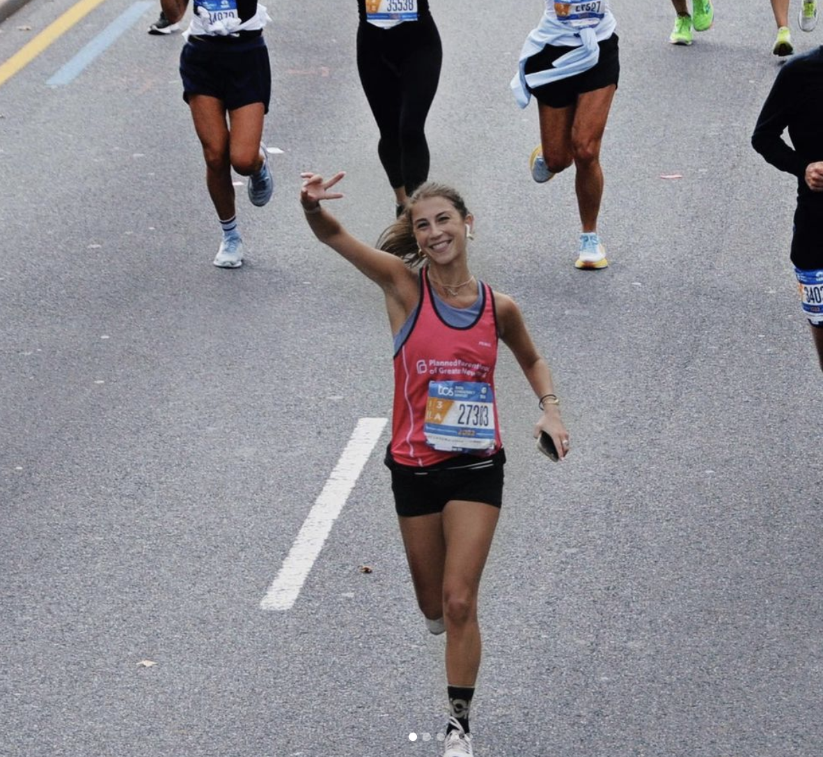 A group of runners participating in a marathon or race, with a woman in a red top and black shorts smiling and making a peace sign while running.