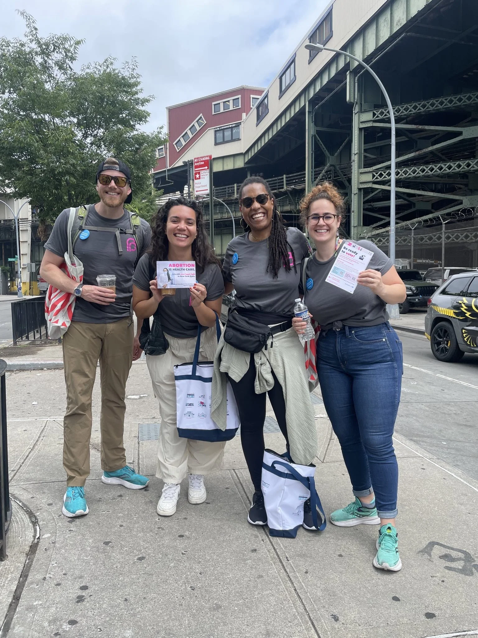 Group of four people wearing matching T-shirts, holding flyers, standing on a city sidewalk with an elevated train track in the background.