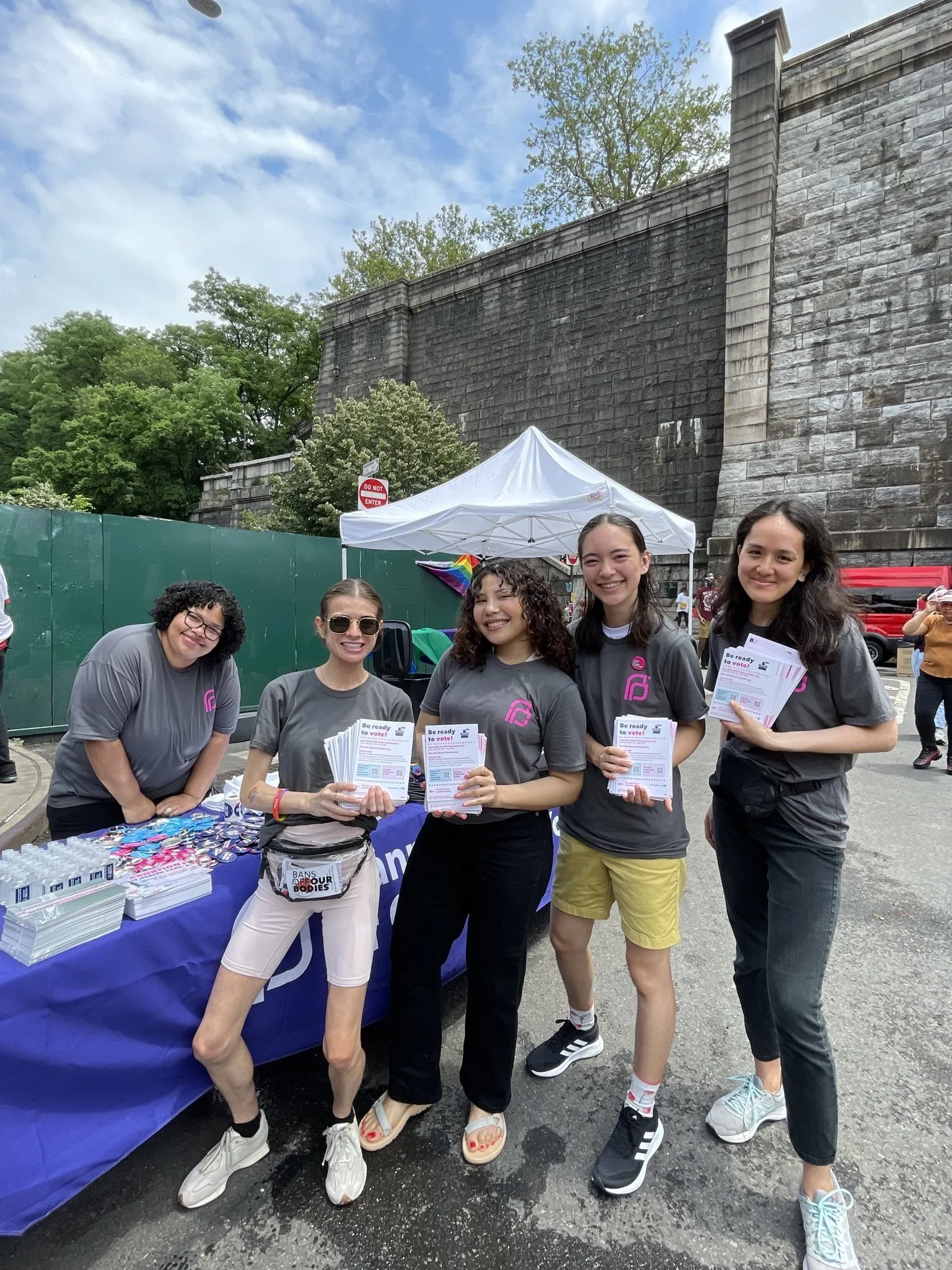 Group of people standing by a table outside, wearing matching gray T-shirts, holding pamphlets, with a tent and stone wall in the background.