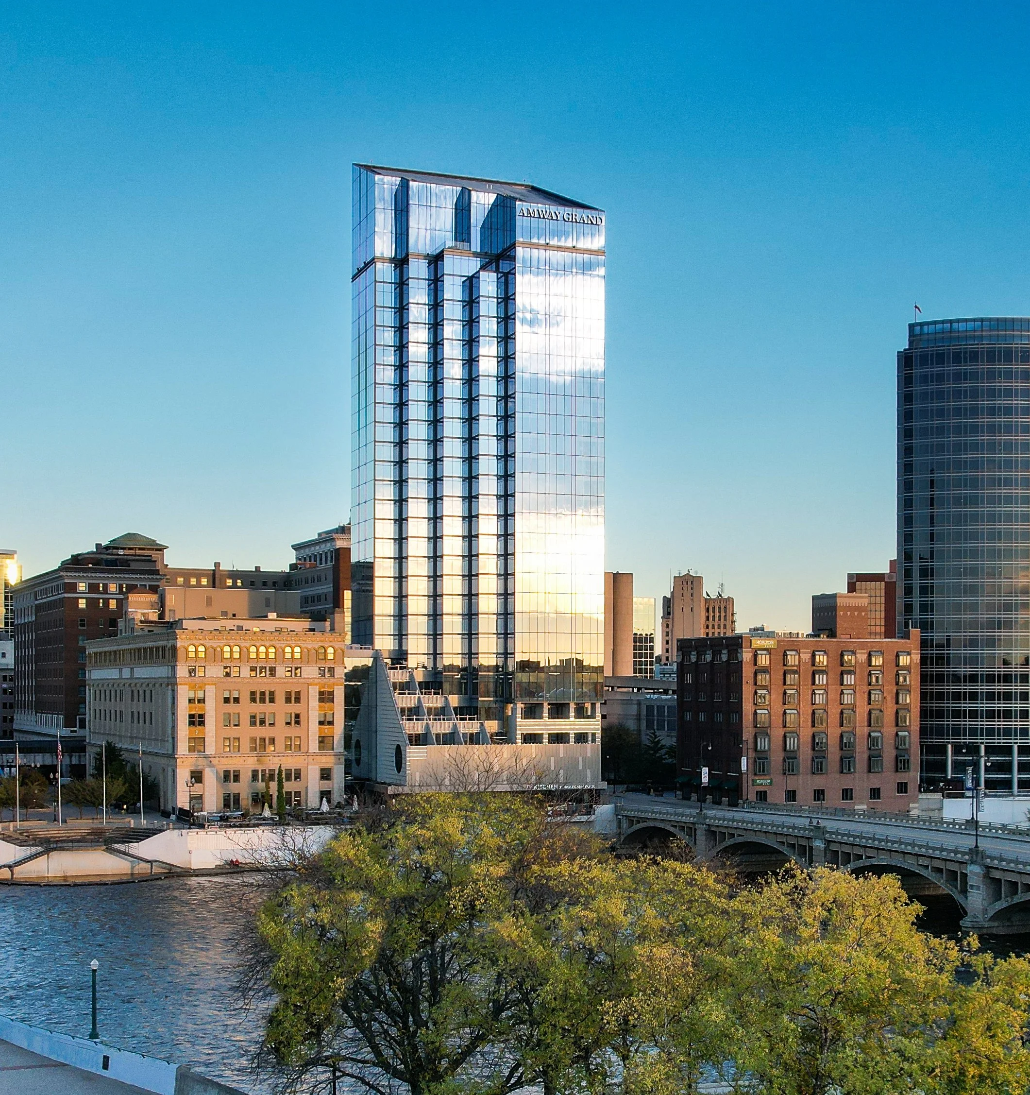 Cityscape featuring a tall glass skyscraper reflecting sunlight, with surrounding buildings and a river, trees, and a bridge in the foreground.