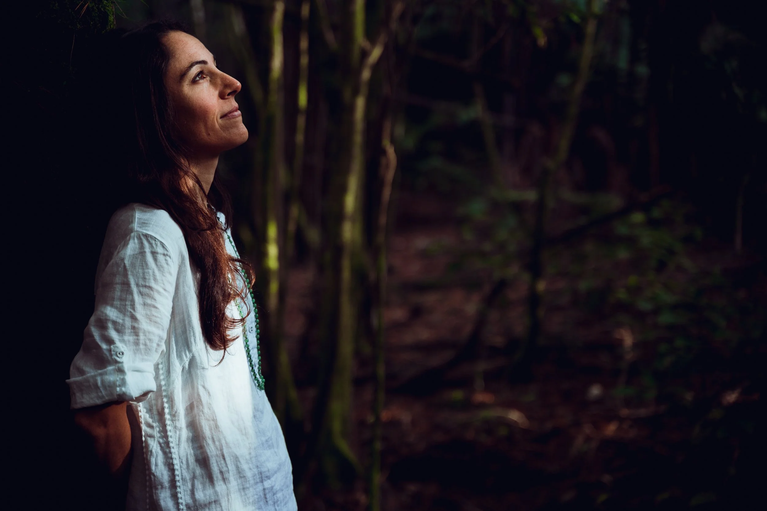 A woman with long brown hair wearing a white shirt and green necklace standing against a dark forest background.