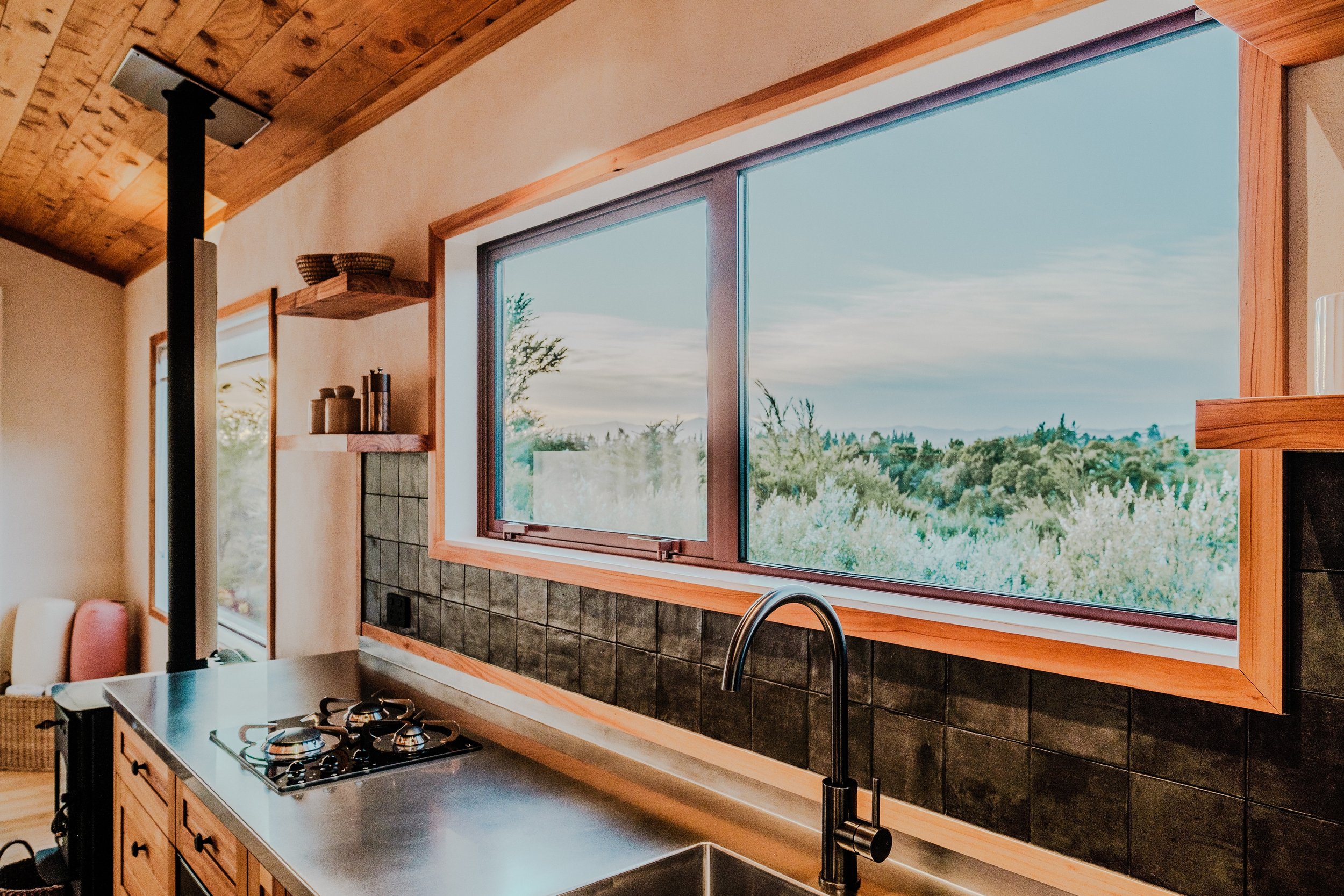 Kitchen with a large window overlooking a lush green landscape, wooden cabinets, a stainless steel countertop with a stove, and a black faucet at the sink.