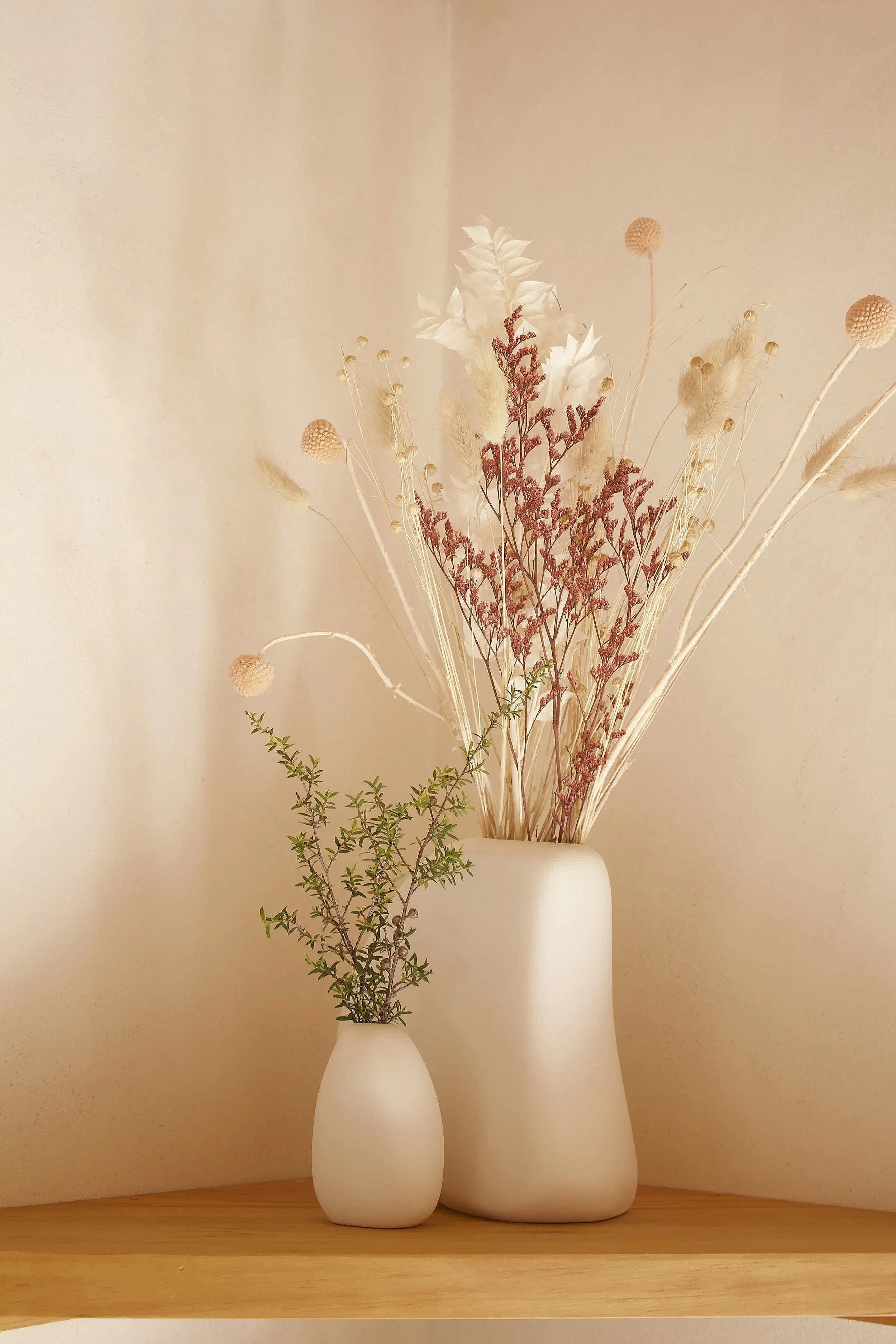 Two white ceramic vases holding dried flowers and plants, placed on a wooden surface against a light beige wall.
