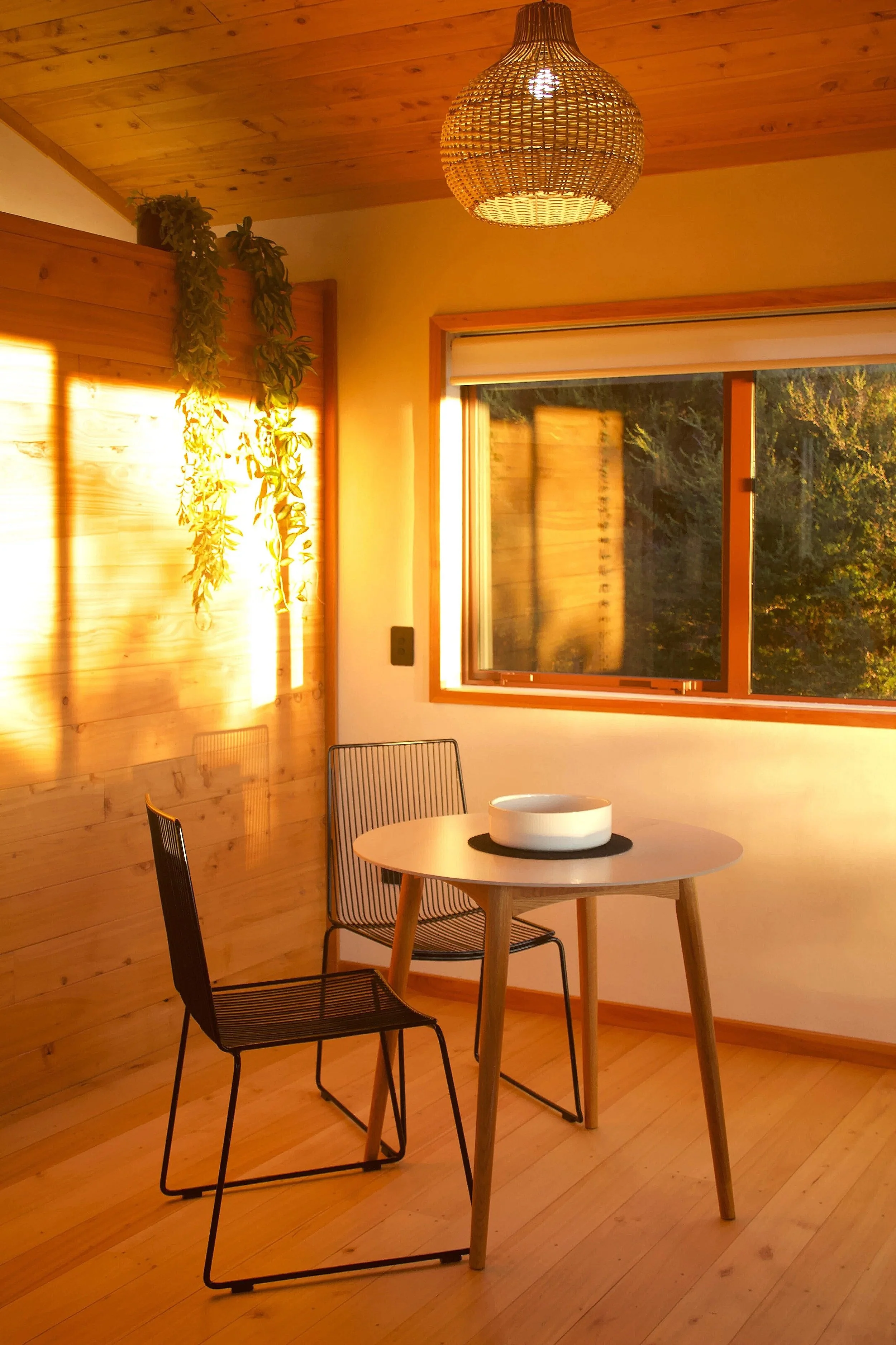 A cozy dining area with wooden flooring and walls, a small round wooden table, two black metal chairs, a white bowl on a black placemat, a window with a view of trees, and a woven pendant light hanging from the wooden ceiling.