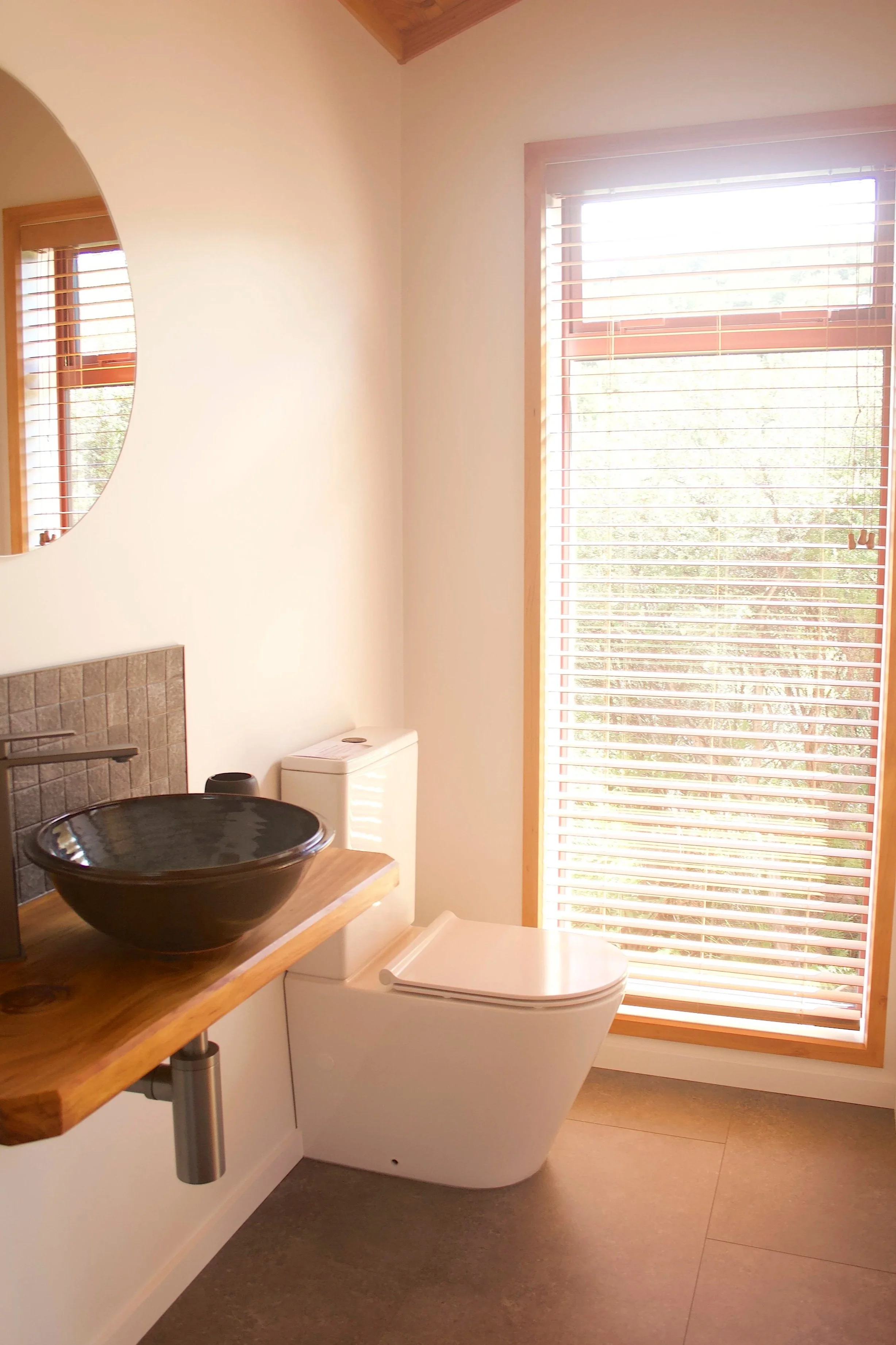 A minimalist bathroom with a black ceramic sink on a wooden countertop, a white toilet, a large window with wooden blinds, a round mirror, and neutral-coloured walls and flooring.